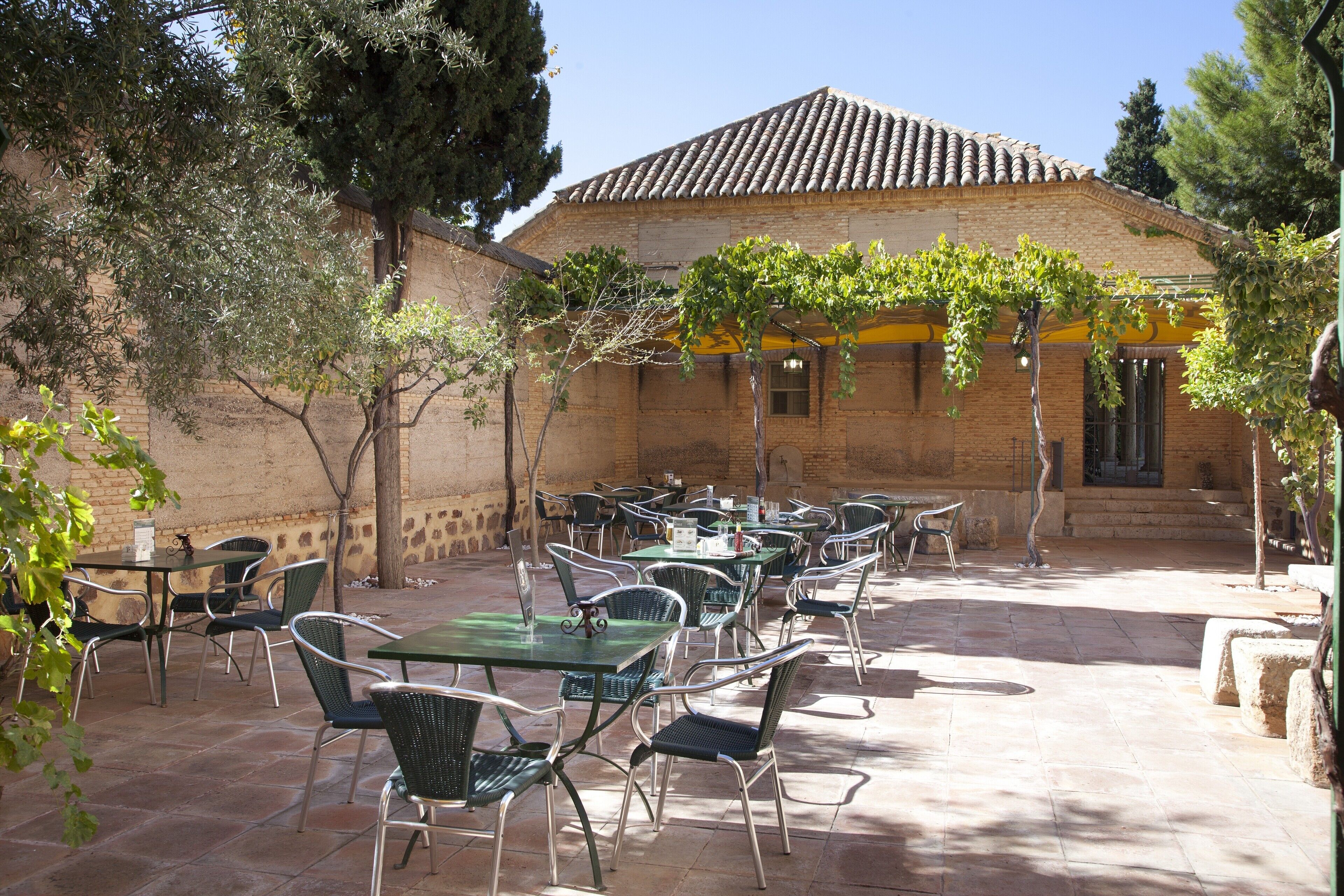Une terrasse extérieure avec des tables et des chaises, un bâtiment en briques avec un toit en tuiles et une pergola couverte de plantes grimpantes.