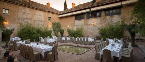 Un área de comedor al aire libre con varias mesas dispuestas para una comida, rodeada de paredes de ladrillo y follaje.