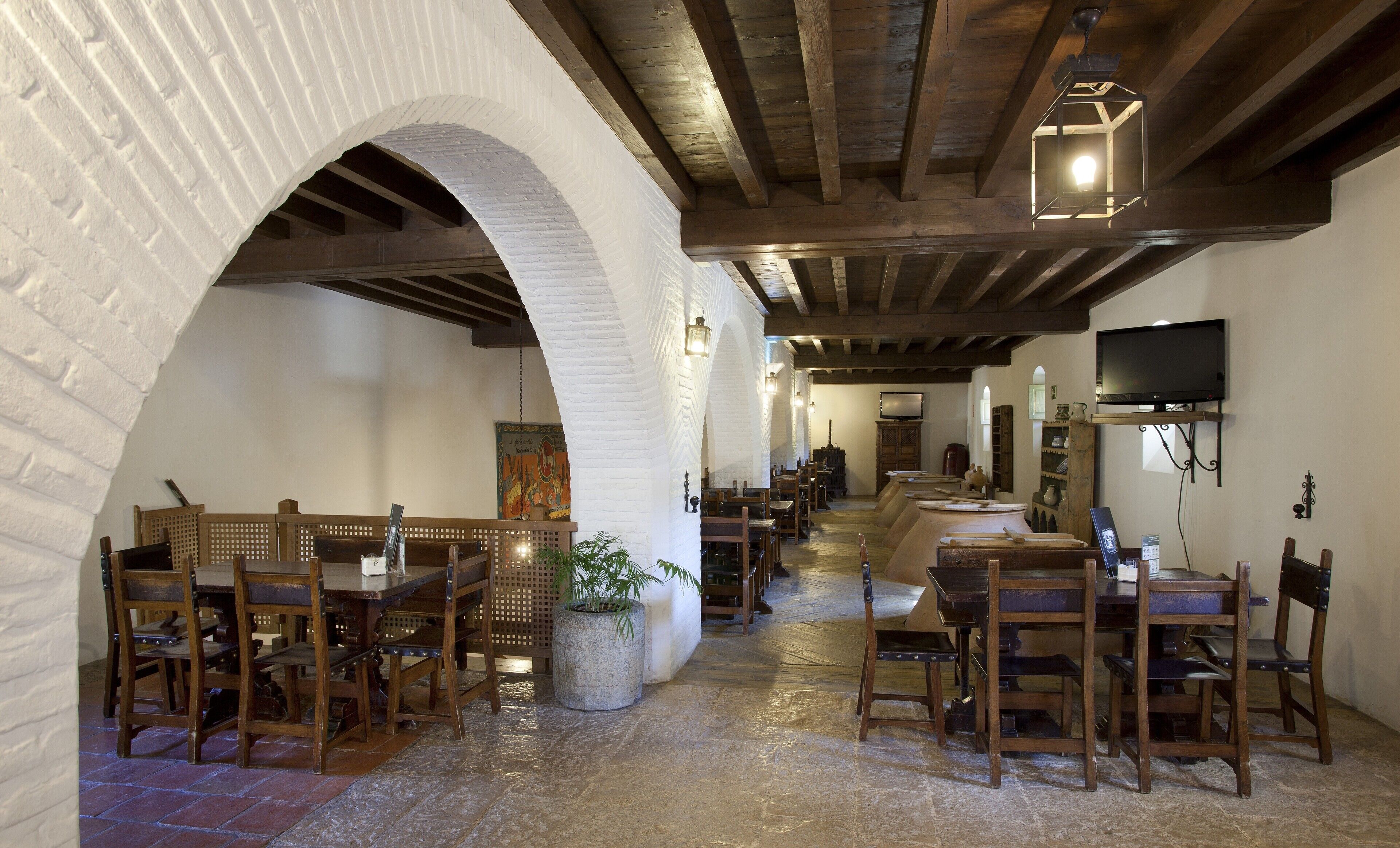 Ein Restaurant mit Backsteinbögen, Holztischen und -stühlen. An der Wand ist ein Fernseher befestigt und in der Mitte steht eine Pflanze.