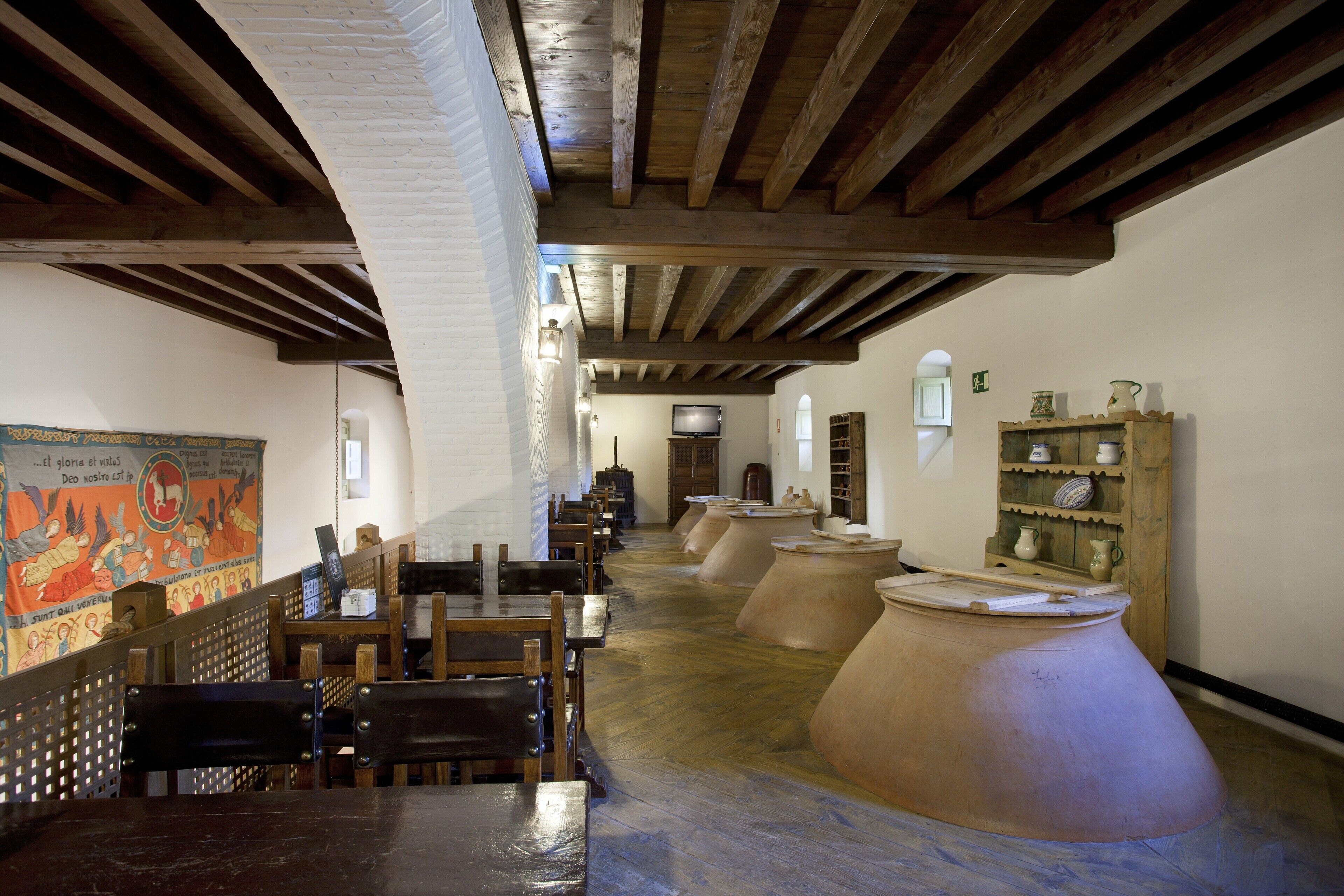 A rustic interior with wooden beams, terracotta pots, and a long table with chairs.