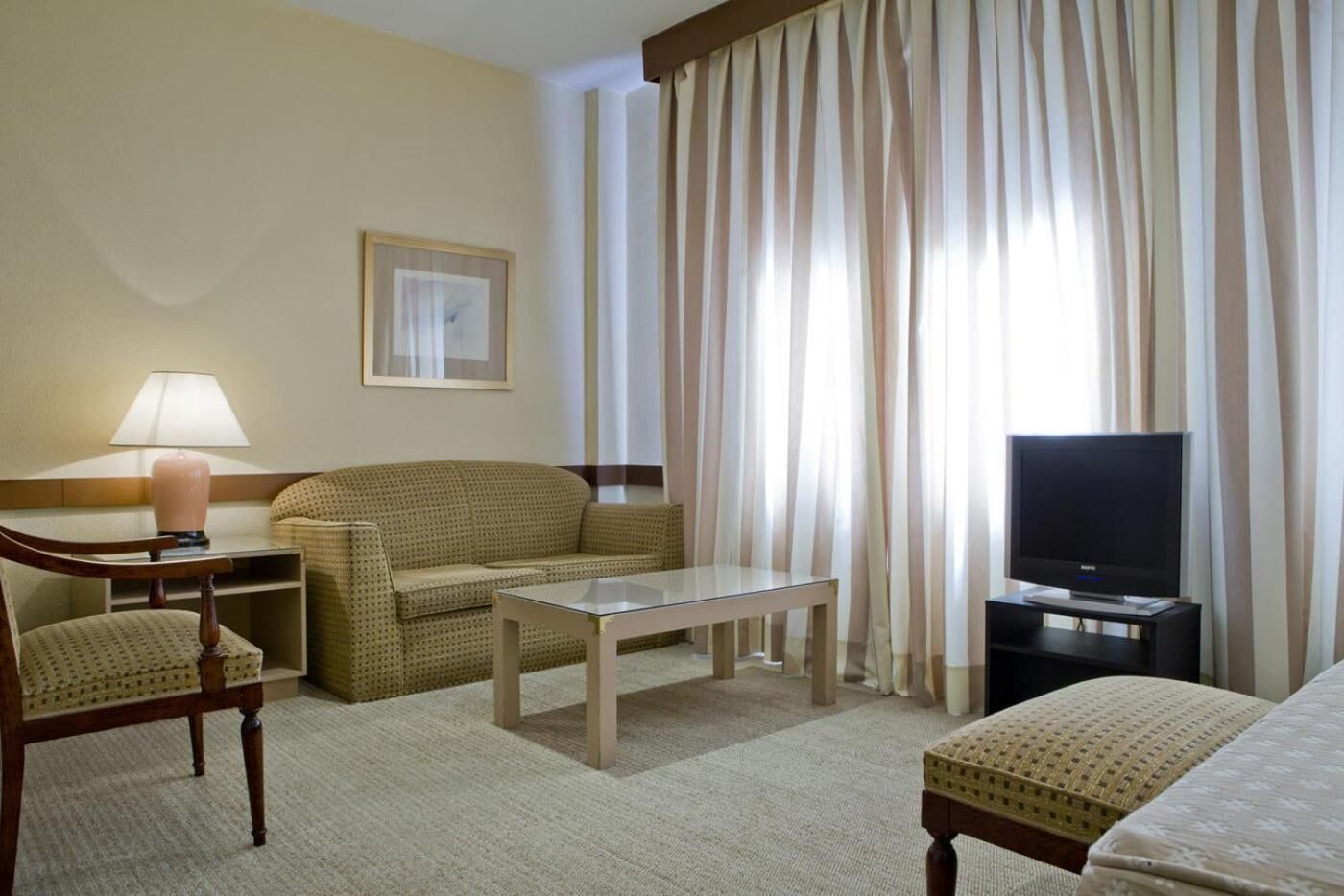 Habitación de hotel con sofá, una mesita, un mueble para televisión, una lámpara y un cuadro en la pared.