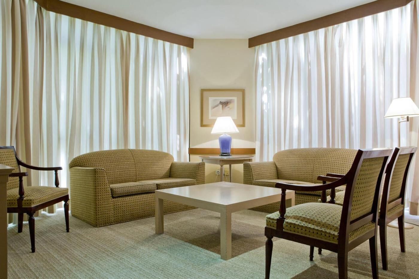 A hotel room with a sofa set, a small table, and two chairs. There is a lamp on a side table and a framed picture on the wall.