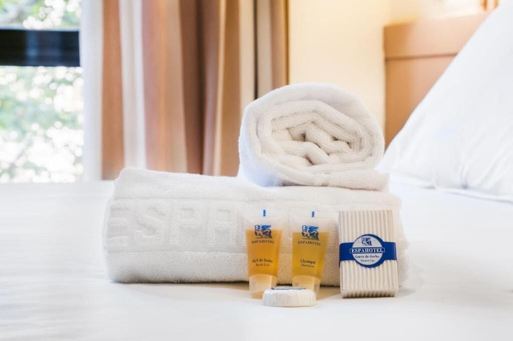 A neatly made bed with a rolled white towel, two bottles of bath gel, and a bar of soap.