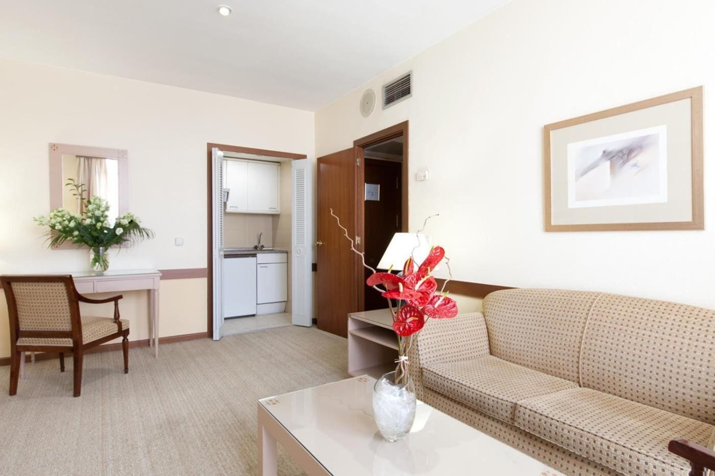 Habitació d'hotel amb un sofà, una taula petita amb un ram, una cadira, escriptori amb un mirall, cuina americana i una porta que dona a una altra habitació.