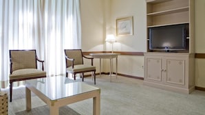 Een hotelkamer met een tafel met glazen blad, twee gestoffeerde fauteuils, een televisie op een kast en een lamp.