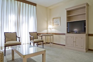 A hotel room with a glass-top table, two upholstered chairs, a television on a cabinet, and a lamp.