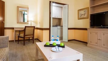 Une chambre d’hôtel avec une table à manger dressée, agrémentée de pommes et de bouteilles d’eau, une chaise, un bureau avec une lampe, un téléviseur et une armoire.