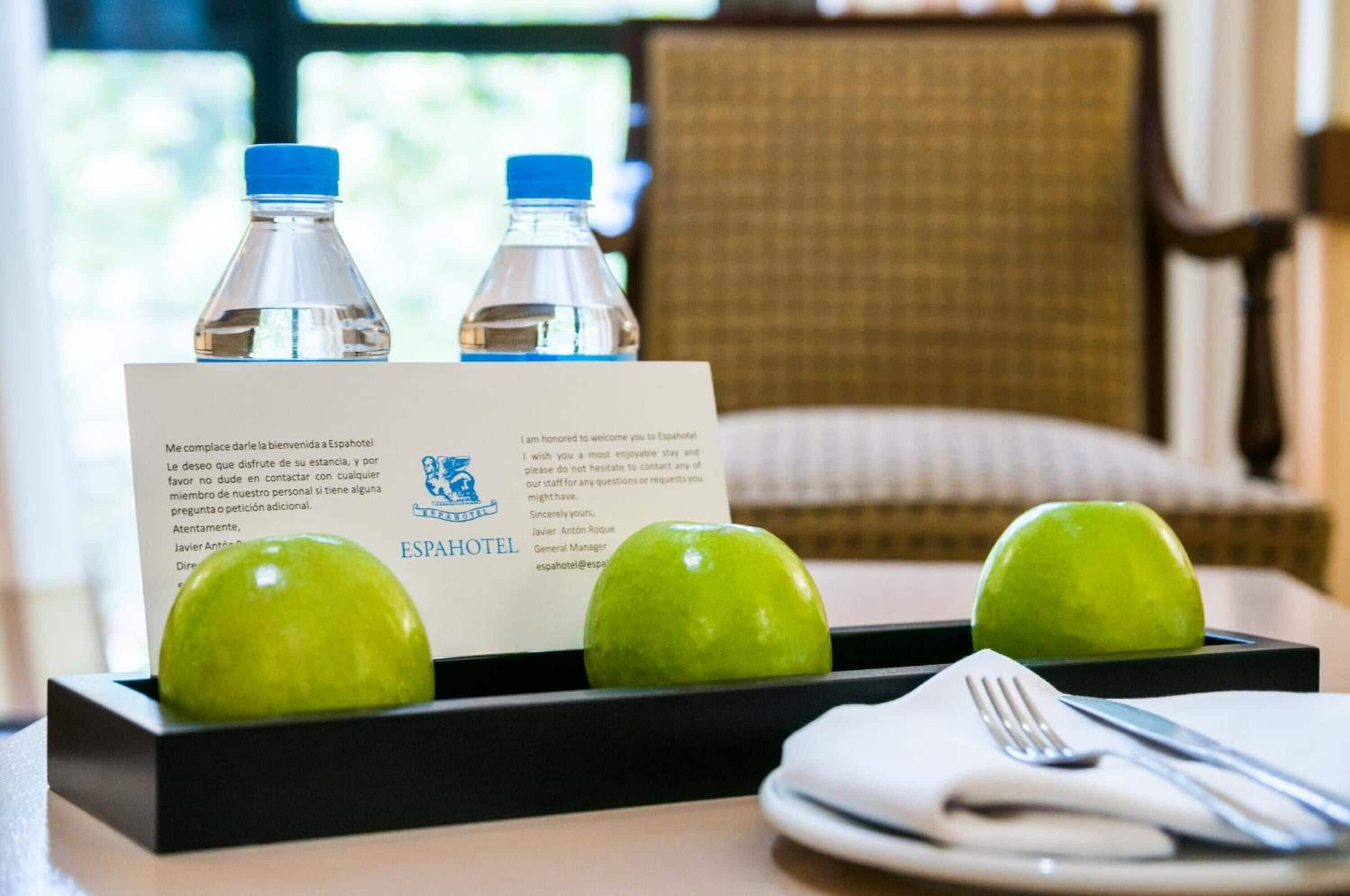 A hotel room with a table set for breakfast, including two water bottles, a plate with two green apples, and a breakfast menu card.