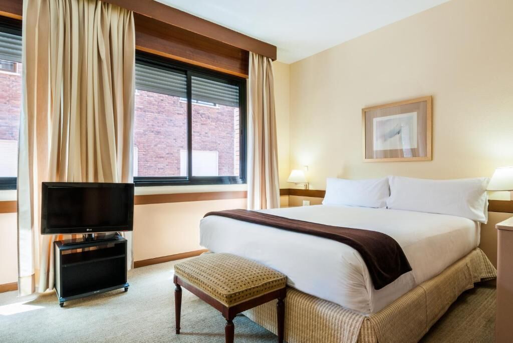 A hotel room with a bed, a TV on a stand, a bedside table, a lamp, and a window with curtains.