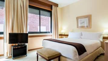Habitación de hotel con cama, televisor sobre un mueble, mesita de noche, lámpara y ventana con cortinas.