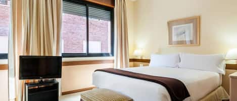 A hotel room with a bed, a TV on a stand, a bedside table, a lamp, and a window with curtains.