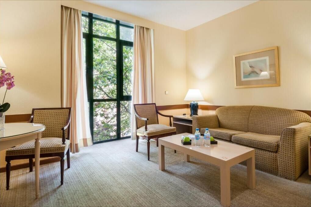 A hotel room with a large window, a sofa, two chairs, a small table, and a lamp.