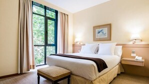 Een hotelkamer met een groot bed, een nachtkastje met een lamp, een klein tafeltje met een stoel, en een raam met gordijnen.