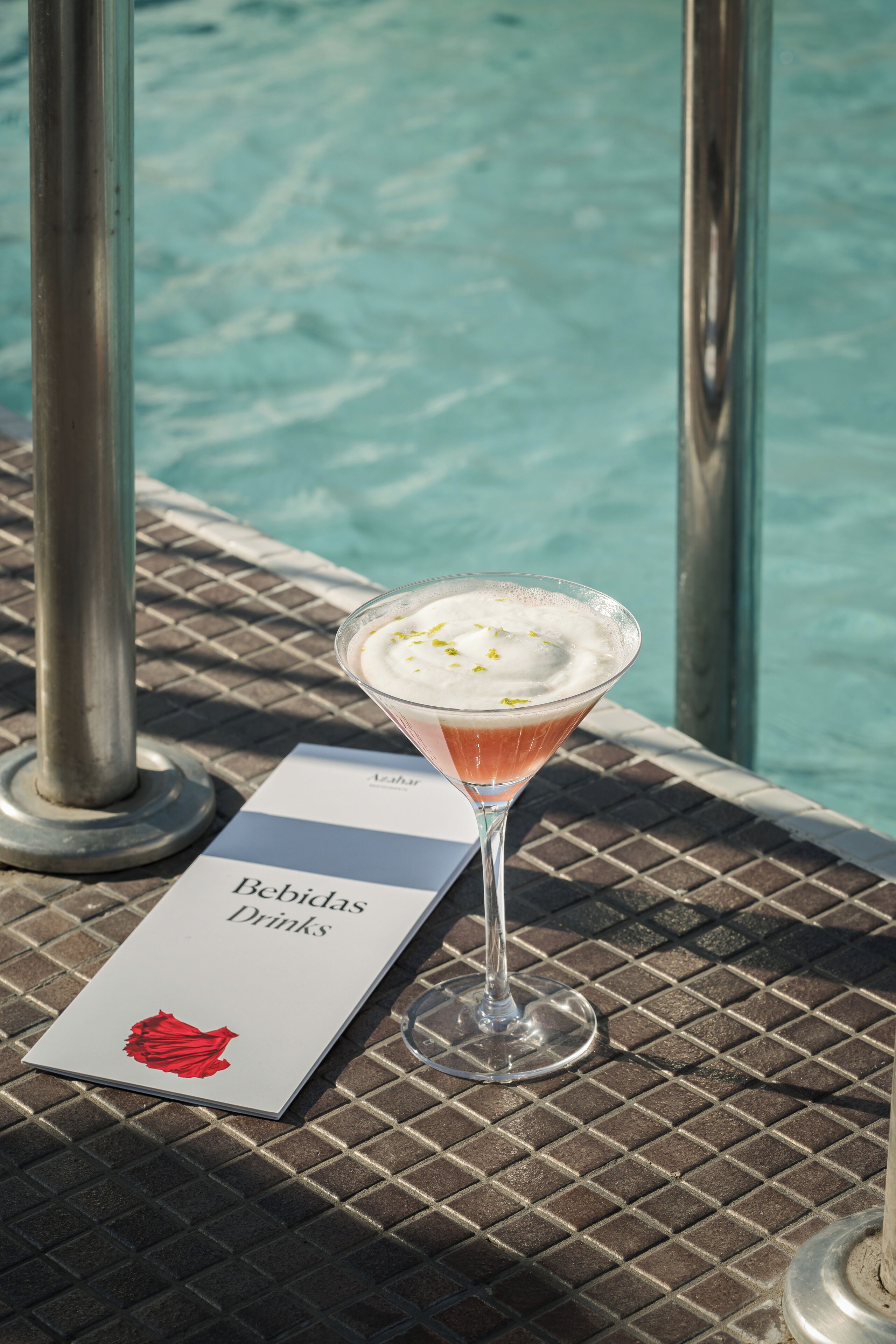 Un verre à martini contenant une boisson rose, posé sur une table en métal au bord d’une piscine.