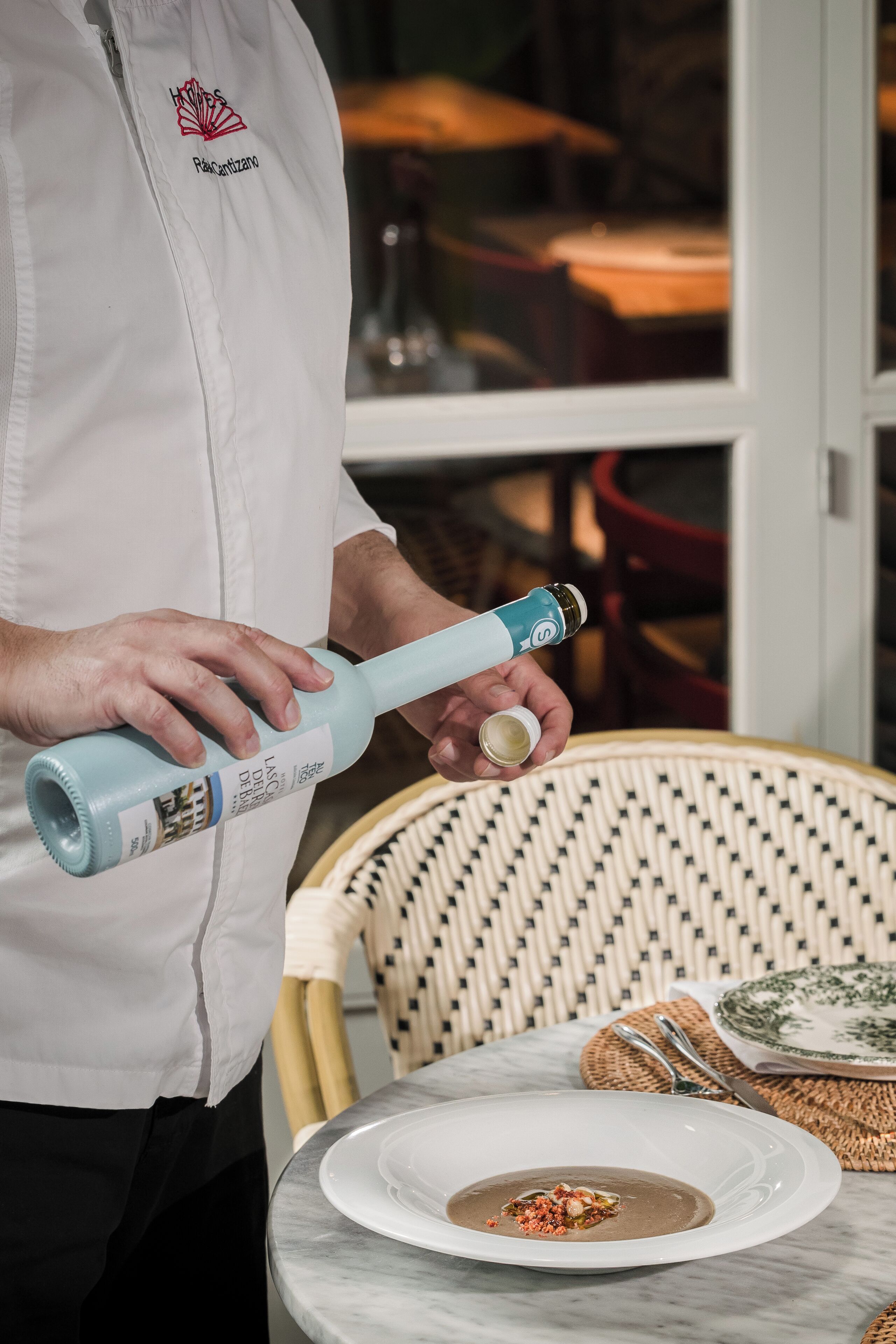 A person in a white uniform with a logo, holding a blue bottle with a white cap, standing next to a table with a white plate and a bowl of soup.