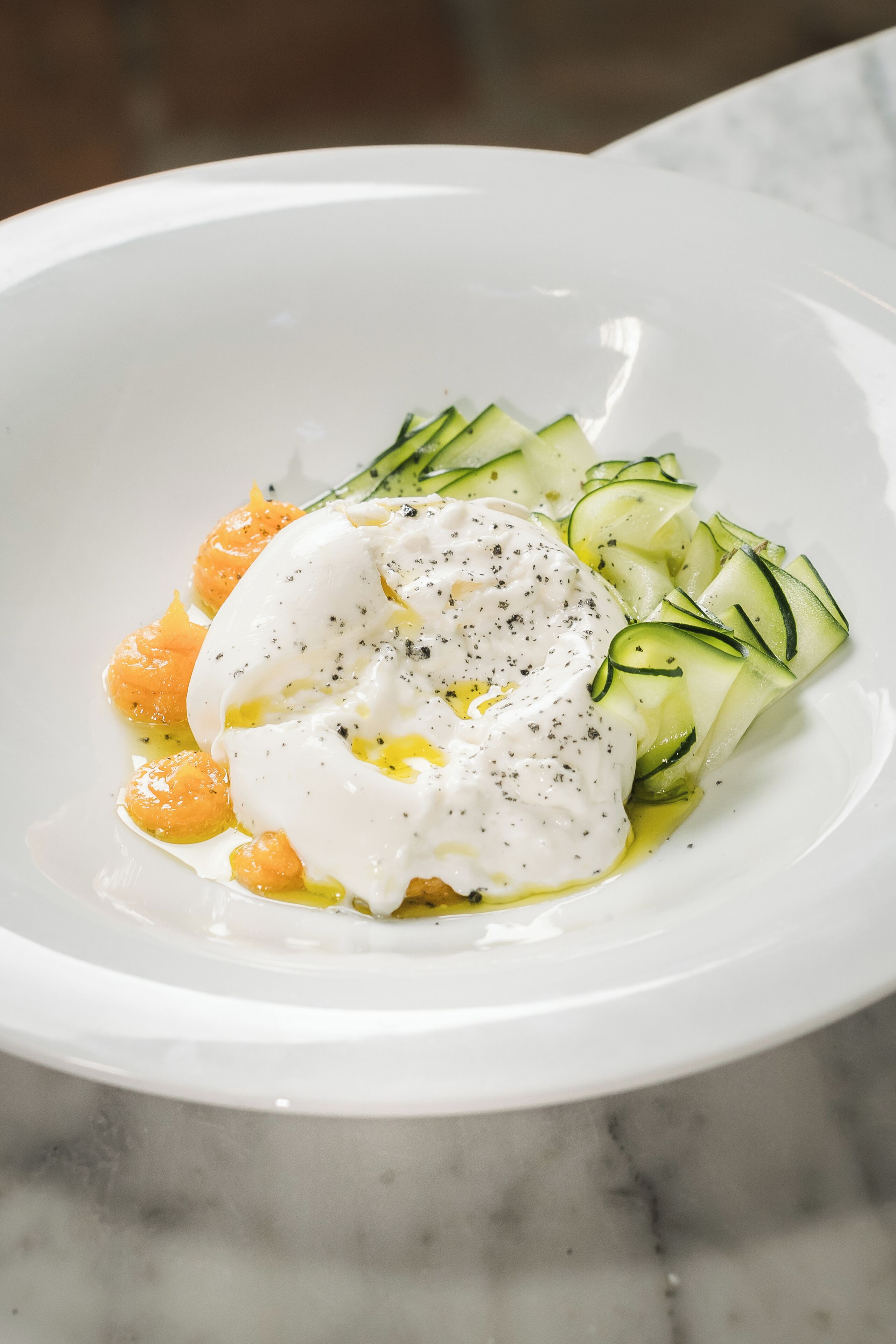 Un bol blanc contenant une assiette avec des œufs pochés, des tranches de courgettes et des segments d’orange.