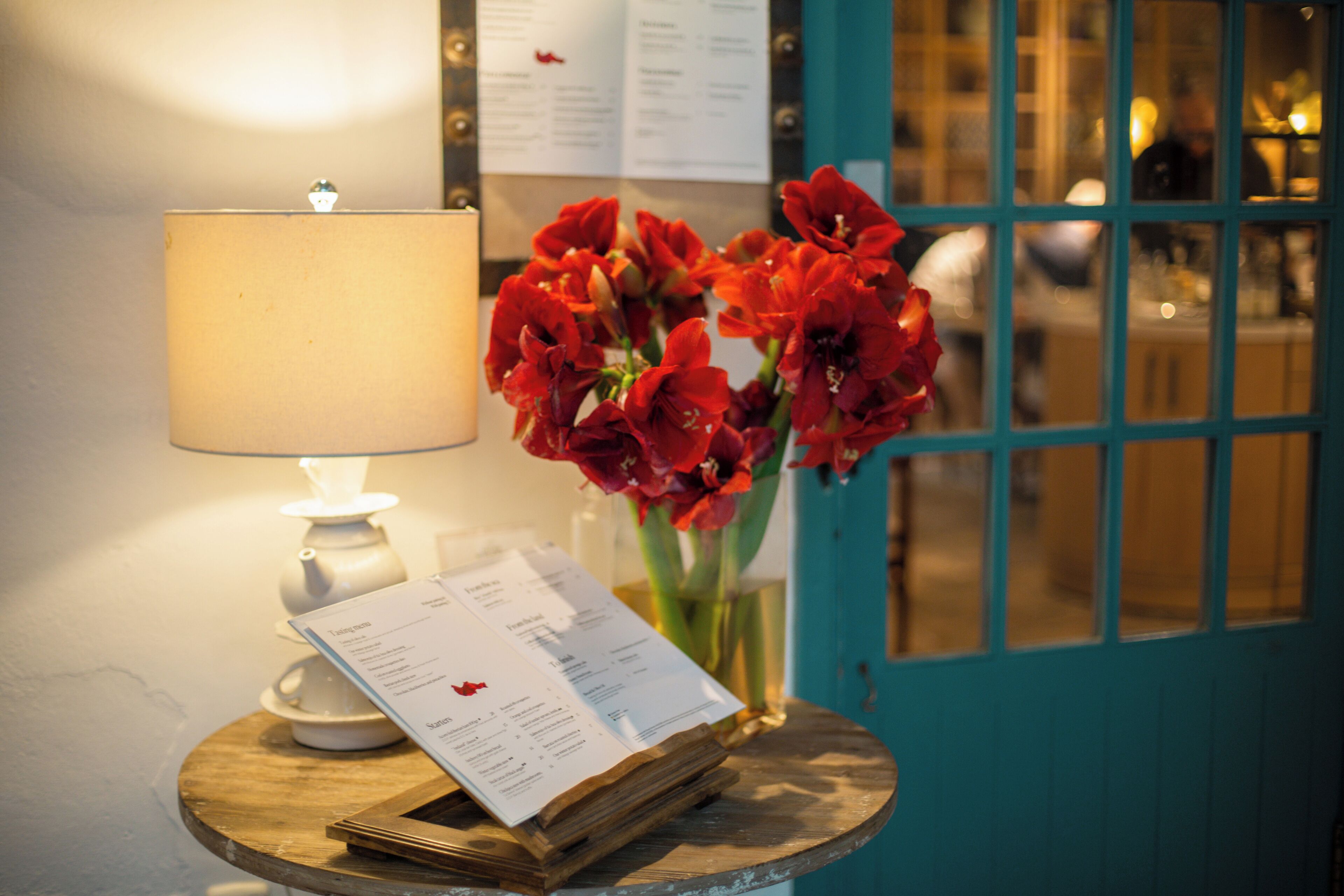 A table with a lamp, menu, and a vase of red flowers.