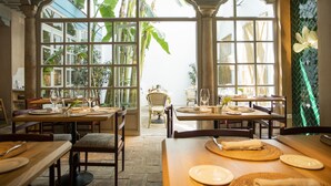 A dining area with tables set for two, large windows, and tropical plants visible through the windows. - Hospes Las Casas del Rey de Baeza, a Member of Design Hotels (Seville)