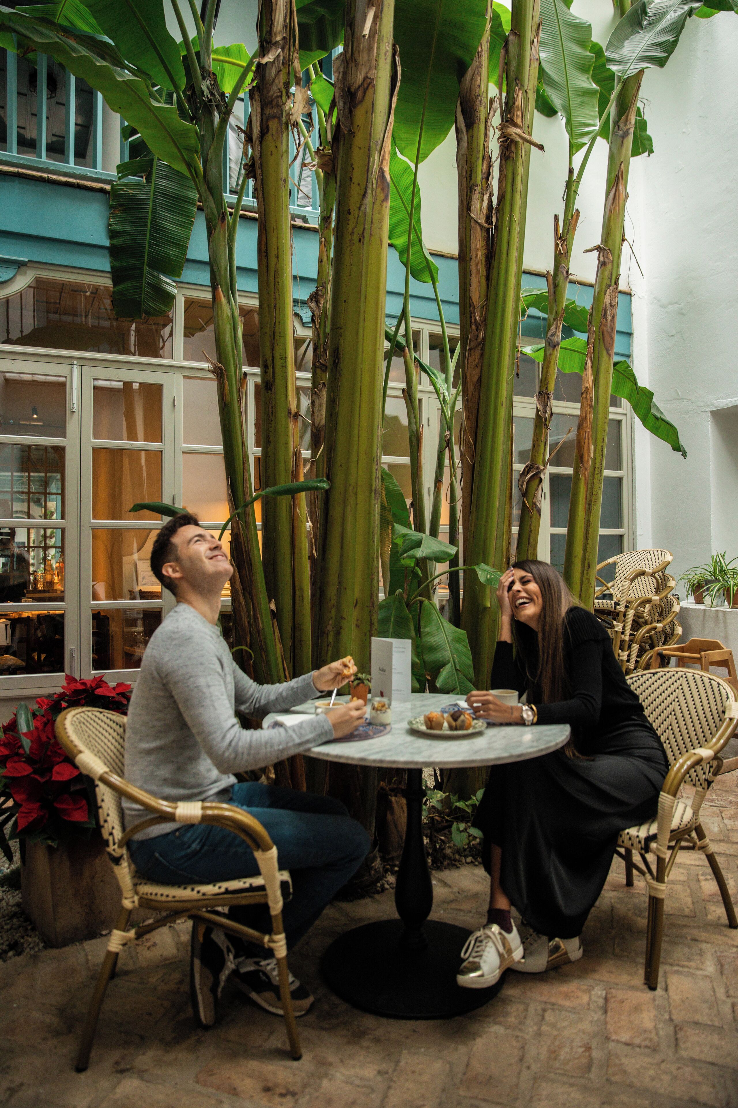 Una pareja cenando en una mesa redonda rodeada de plantas grandes en un ambiente interior.