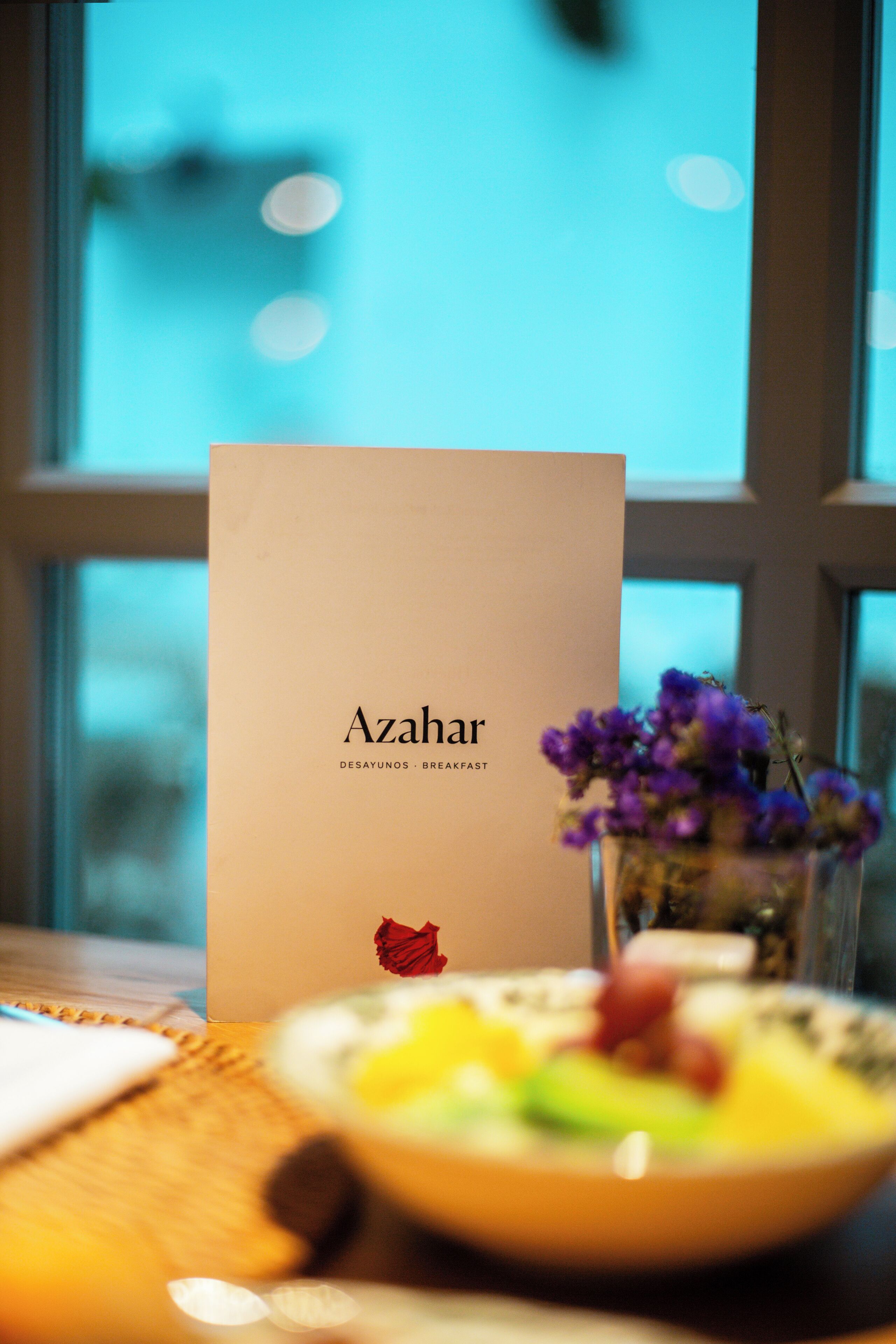 Eine Speisekarte mit dem Titel „Azahar“, eine Schüssel Essen und eine Blumenvase auf einem Tisch neben einem Fenster.