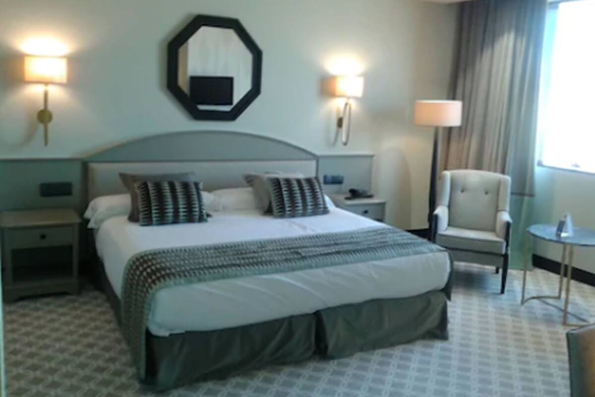 Una habitación de hotel con una cama grande, dos mesitas de noche, una silla, una lámpara y vistas al exterior.