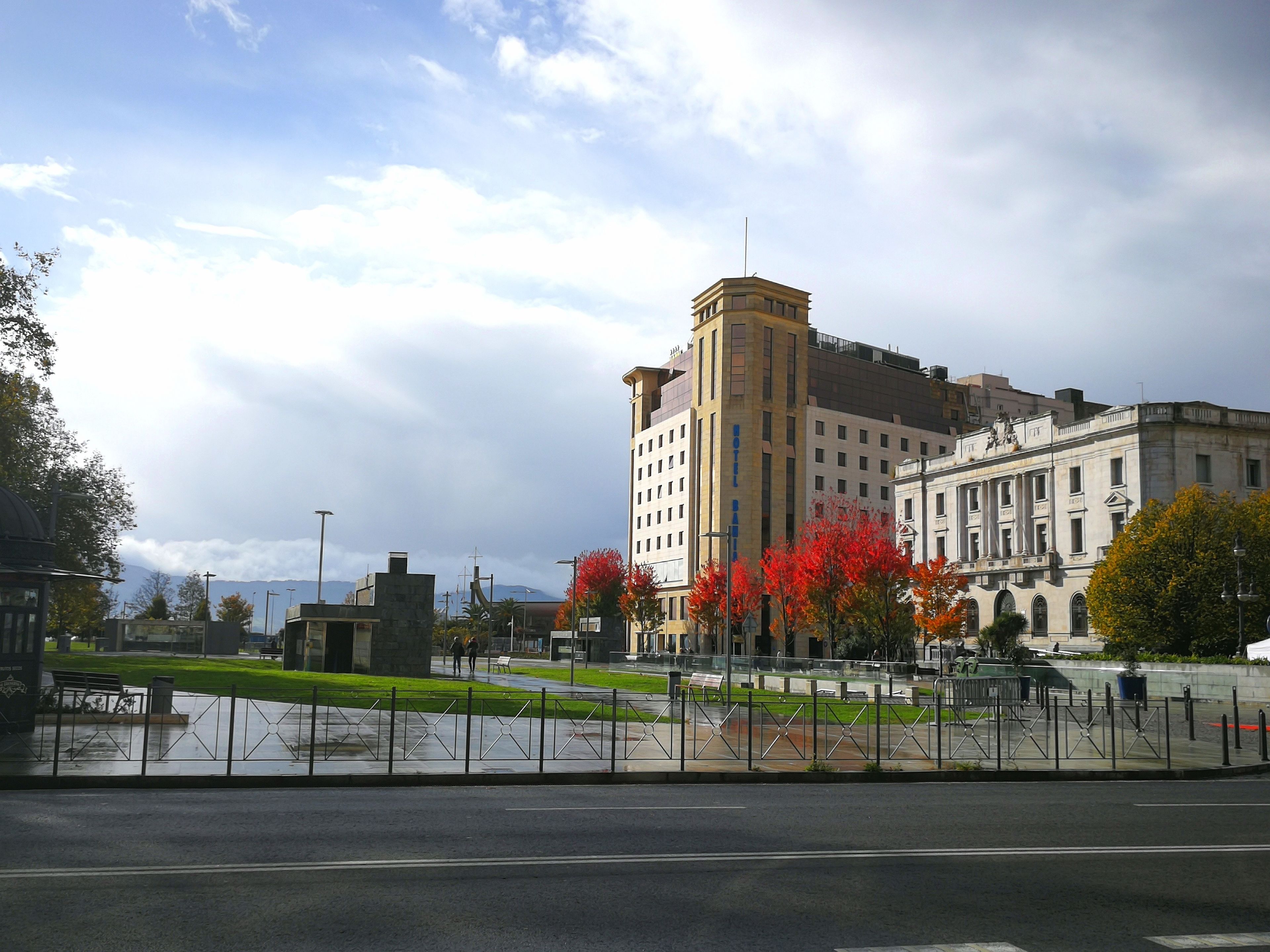A hotel with a central tower, a large building with columns, and a fenced courtyard with trees.