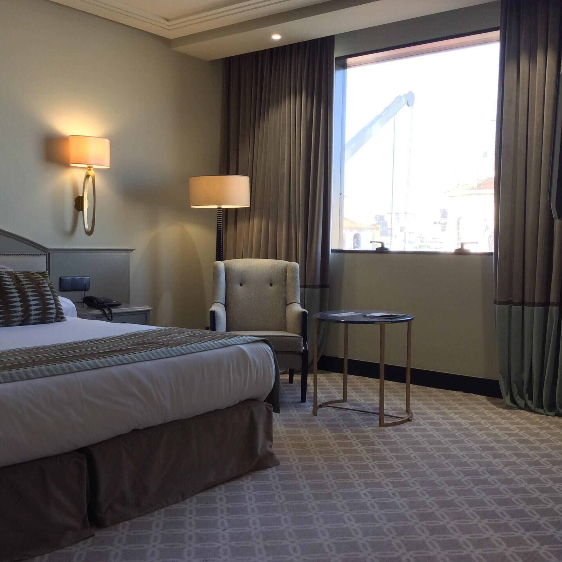 Une chambre d’hôtel avec un lit, une chaise, une petite table, une lampe et une fenêtre avec des rideaux.