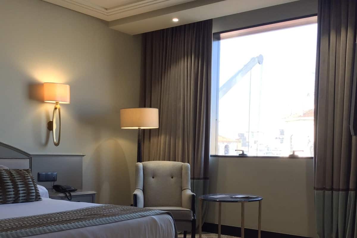 A hotel room with a bed, a chair, a small table, a lamp, and a window with curtains.