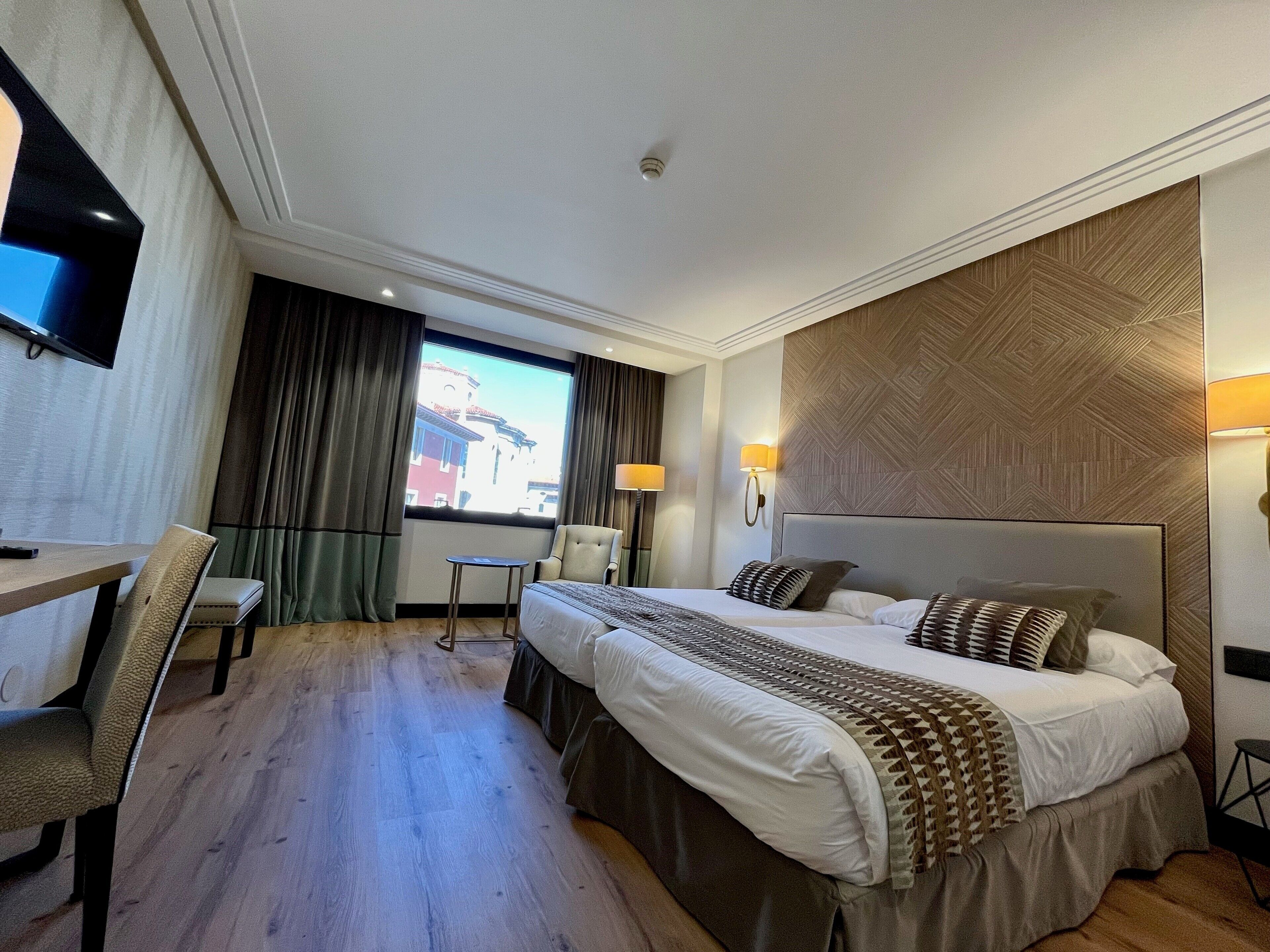 Una habitación de hotel con una cama grande, un escritorio con silla, una mesita pequeña y un televisor fijado a la pared.