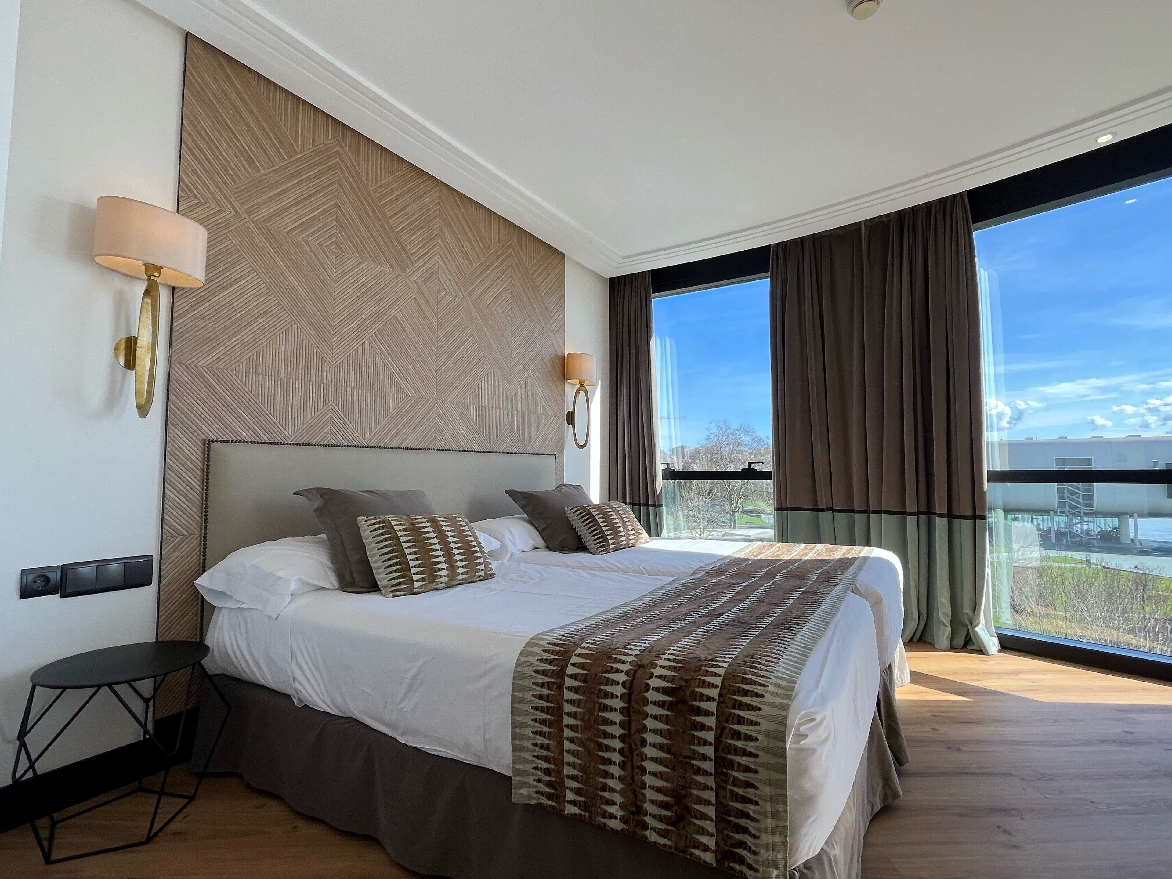 Una habitación de hotel moderna con una cama grande, una mesita y vistas a la ciudad a través de amplios ventanales.