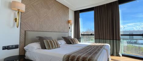 Una habitación de hotel moderna con una cama grande, una mesita y vistas a la ciudad a través de amplios ventanales.