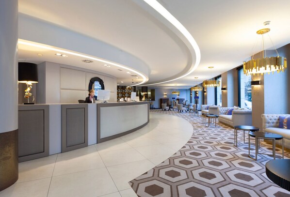 A modern hotel lobby with a curved reception desk, seating area with sofas and tables, and large windows providing natural light.