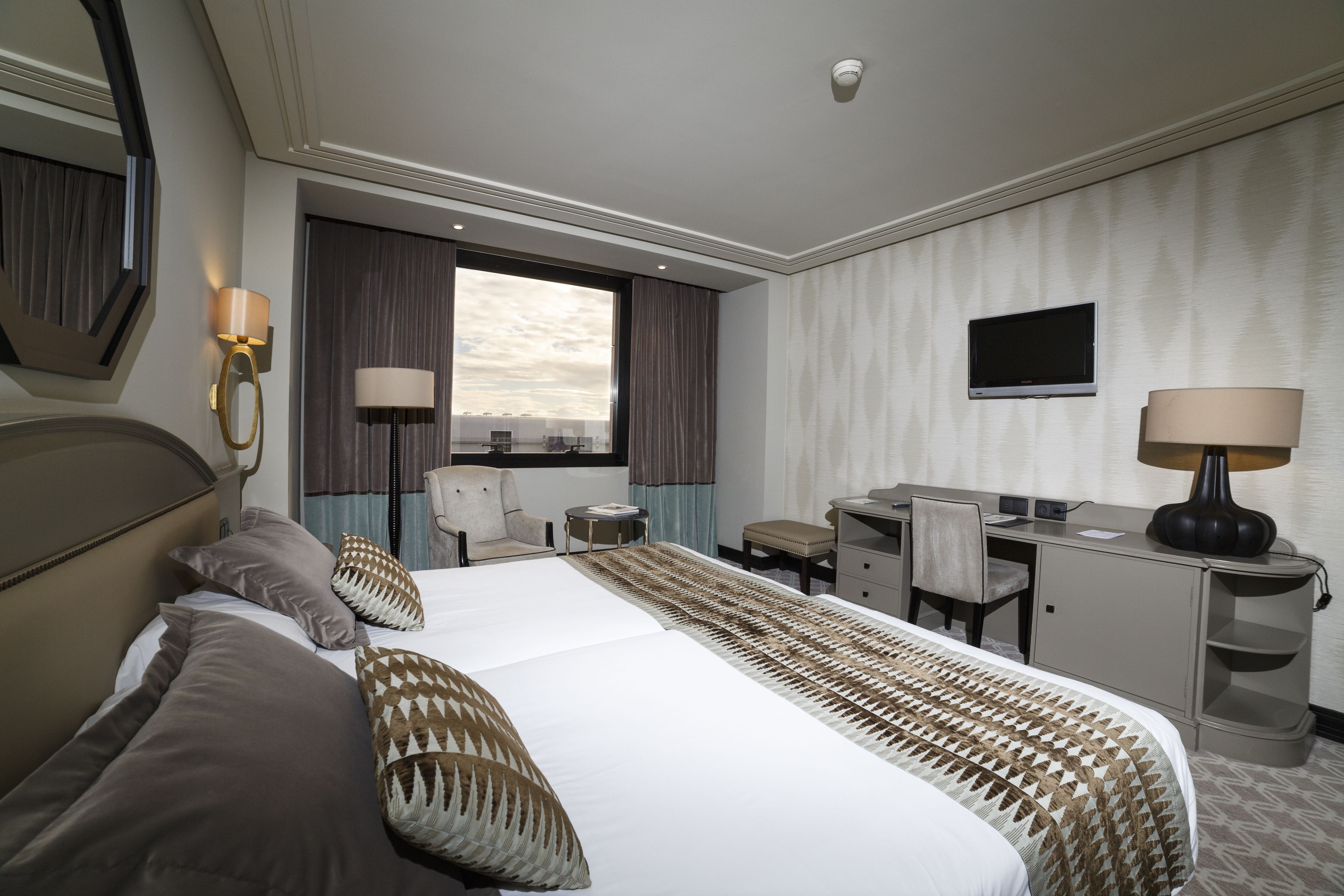 A hotel room with a large bed, a desk, a chair, a TV, and a window with curtains.