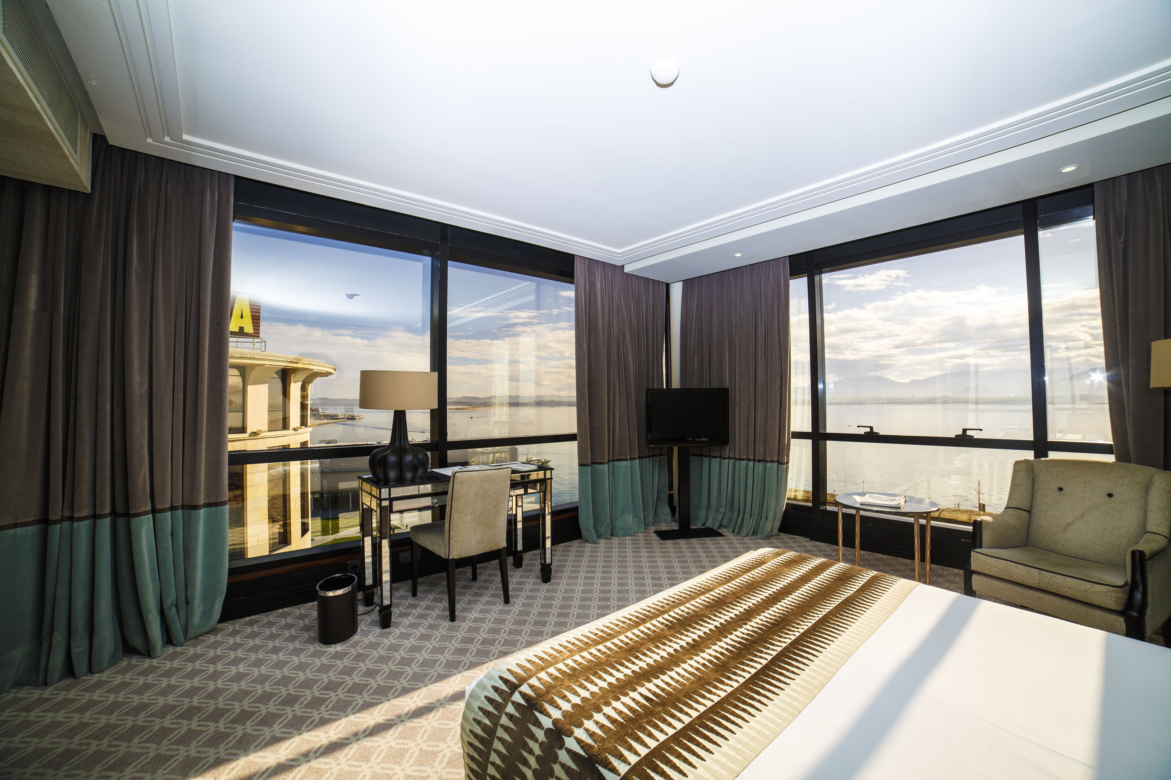 A hotel room with a large bed, a desk with a chair, a TV, and a view of the cityscape through large windows.