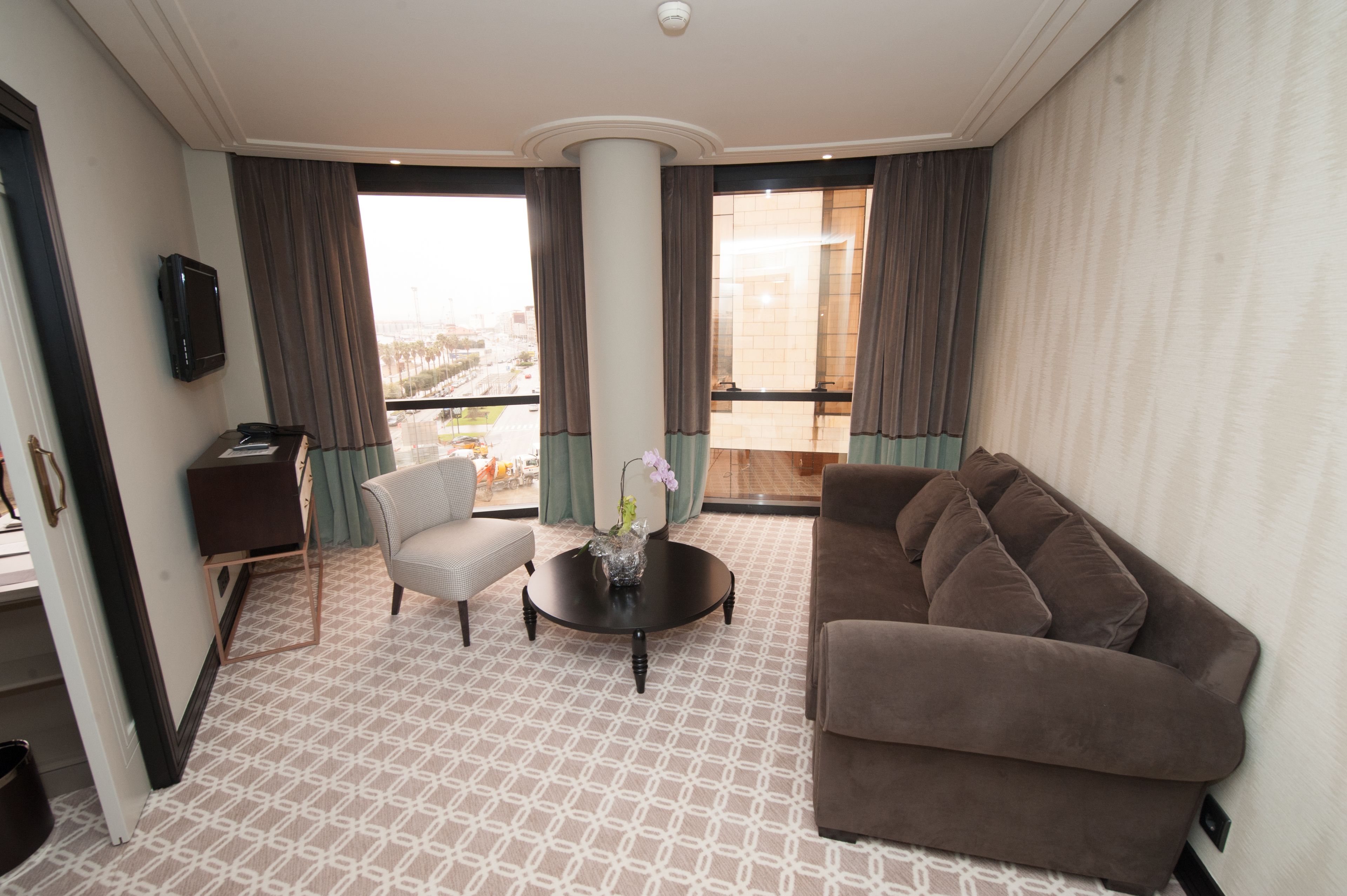 A hotel room with a large window, a brown sofa, a white armchair, a small table with a vase, and a television on the wall.