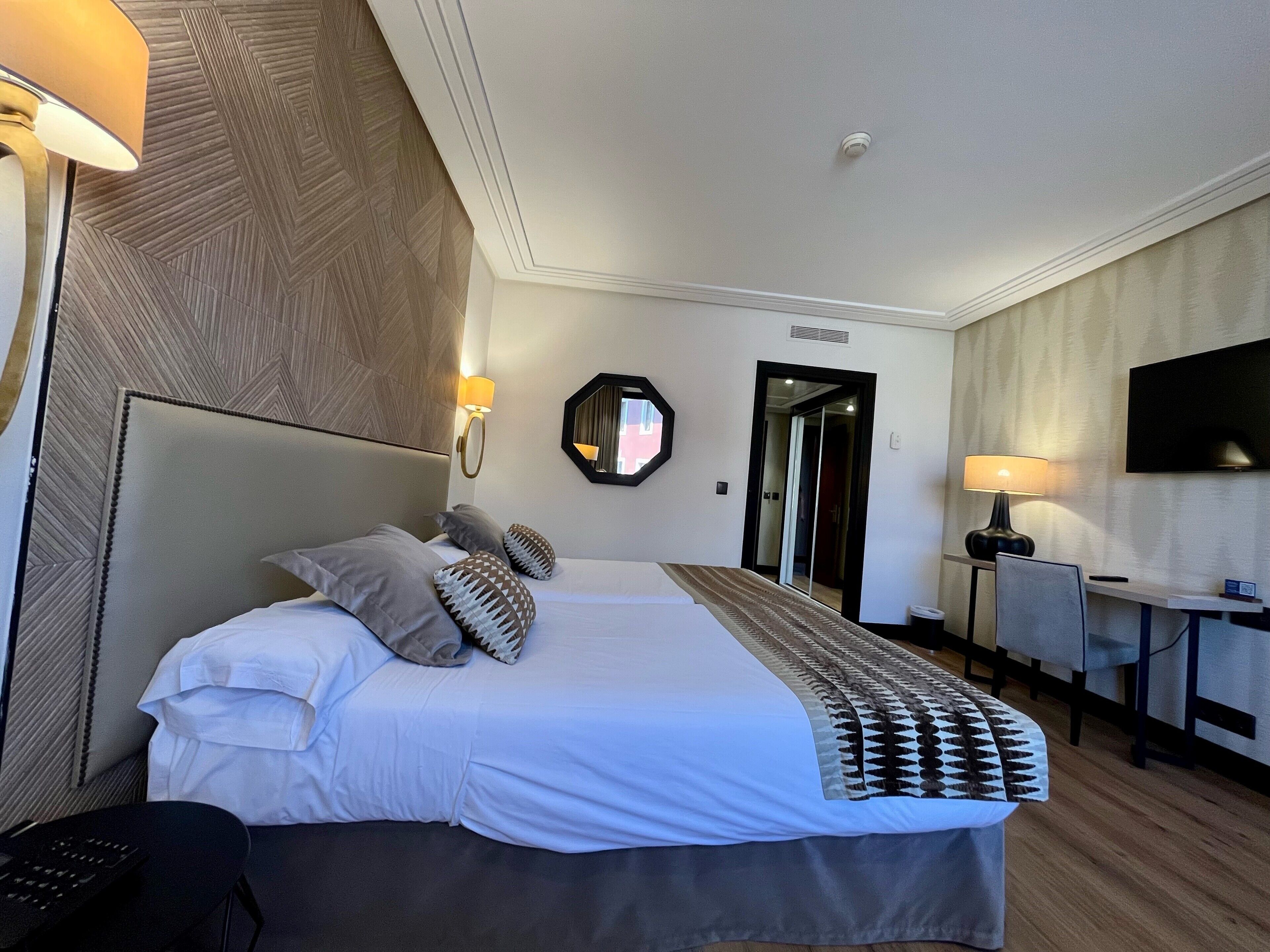 Ein Hotelzimmer mit einem großen Bett, zwei Nachttischlampen, einem Schreibtisch mit Stuhl, einem Flachbildfernseher und einem Spiegel an der Wand.