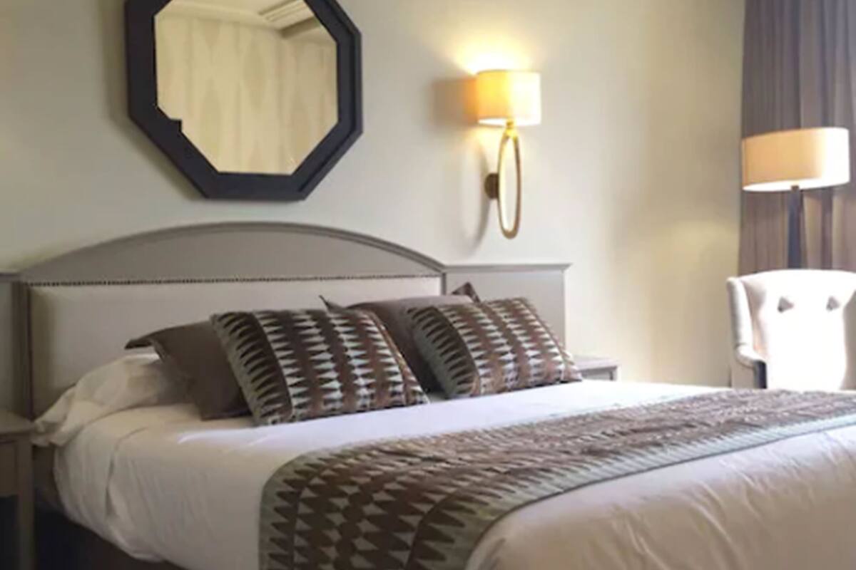 Una cama bien tendida con almohadones estampados, un cabecero y dos lámparas fijadas a la pared.