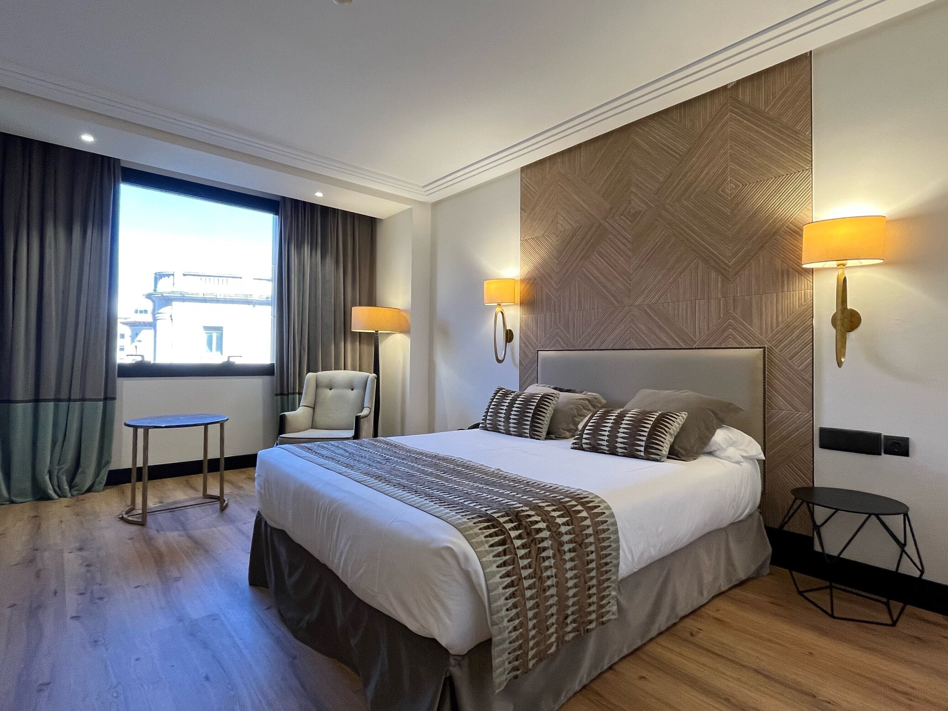 Quarto de hotel moderno com uma cama grande, duas luminárias de cabeceira, uma cadeira e uma mesa pequena.