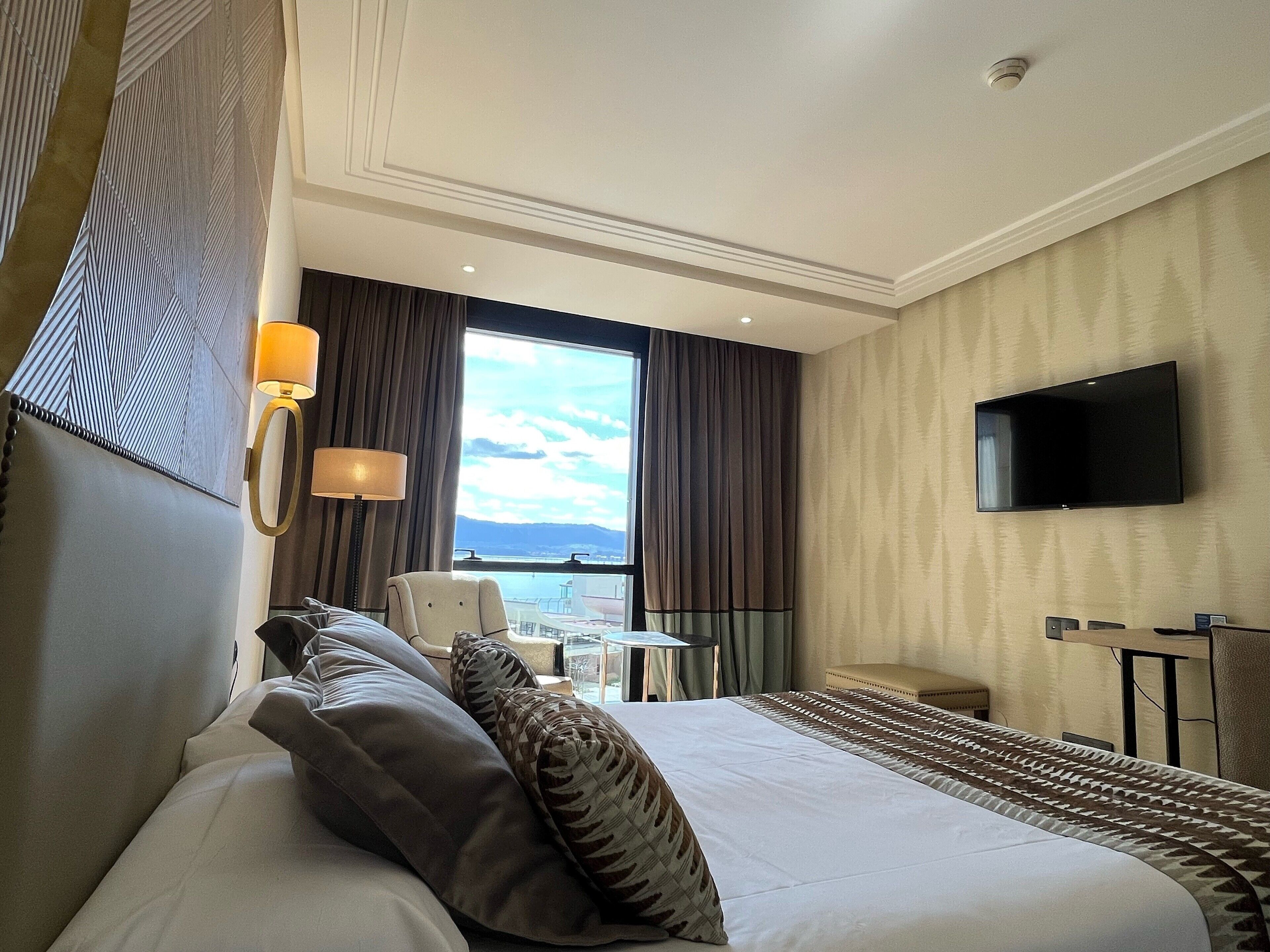 A hotel room with a large bed, a flat-screen TV mounted on the wall, a desk with a chair, and a window with curtains.