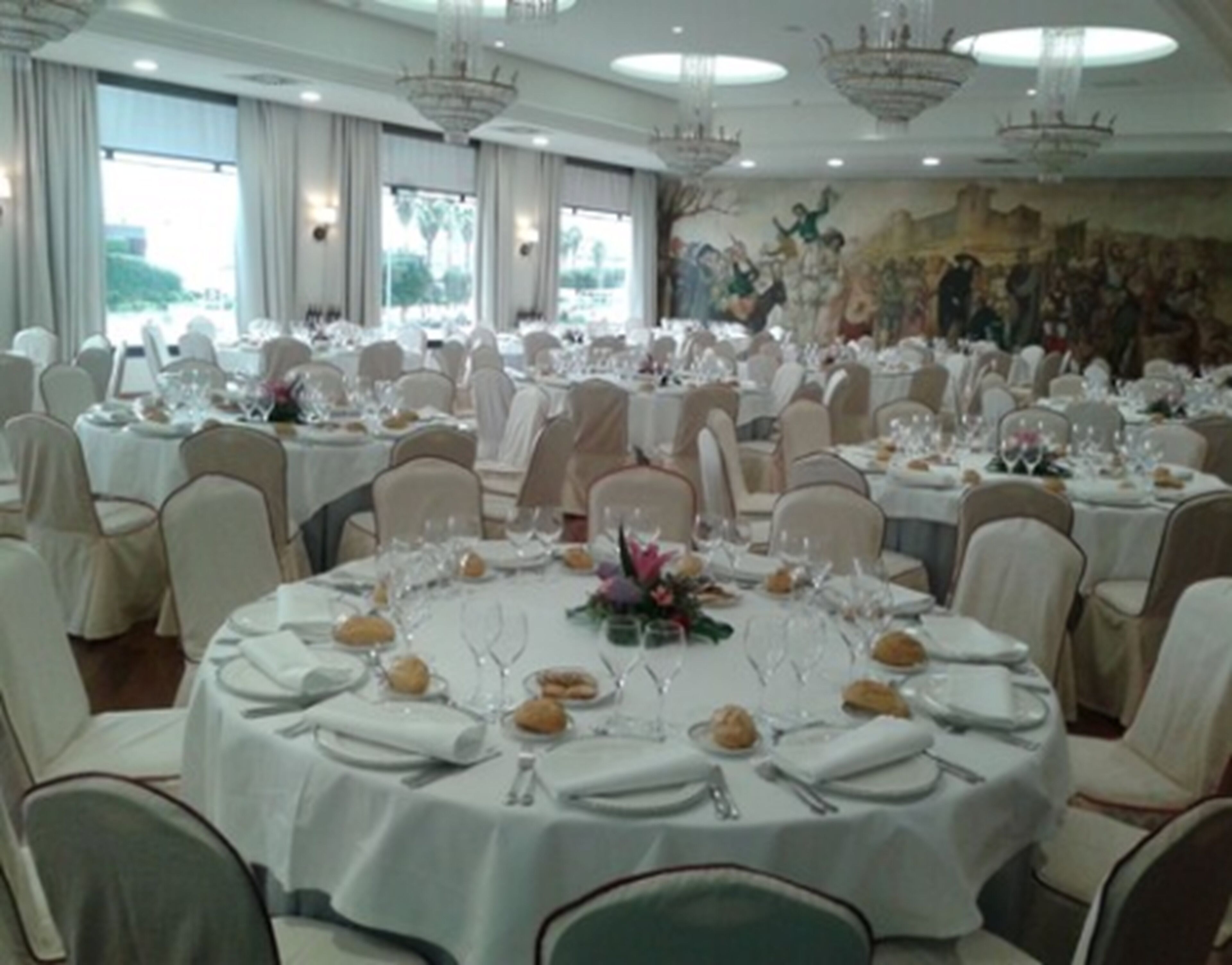 A banquet hall with round tables set for a formal event, featuring chandeliers and a mural on the wall.