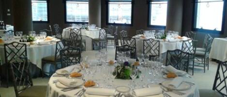 A dining area with round tables set for a meal, featuring white tablecloths and metal chairs.