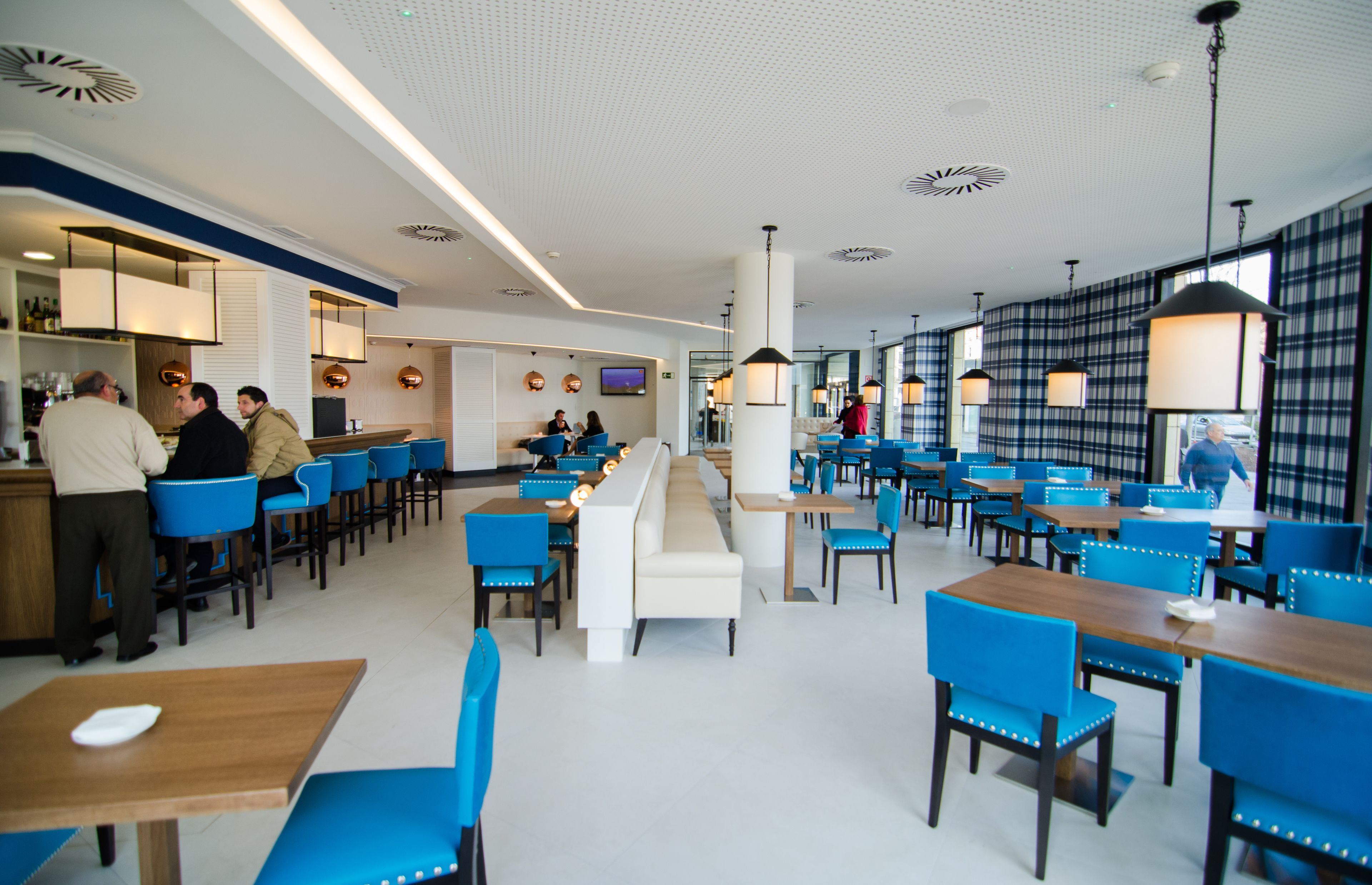 A modern restaurant with blue chairs and wooden tables, a bar area, and a central seating area with a white bench.