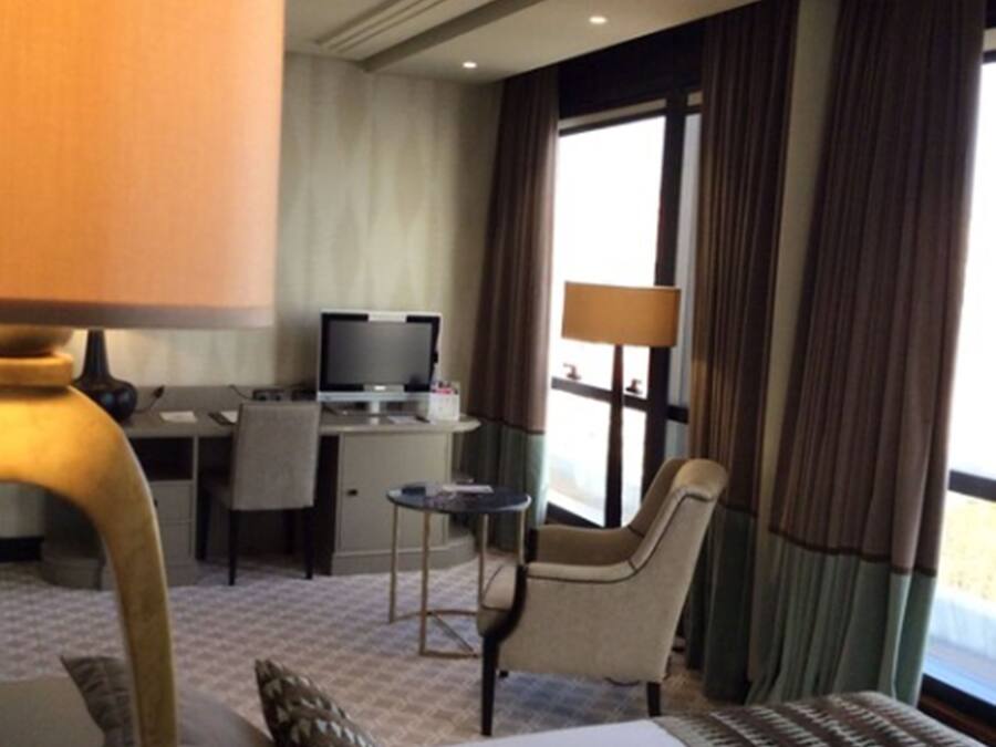 Habitación de hotel con una cama, un escritorio con computadora, una silla, una lámpara y una ventana con cortinas.