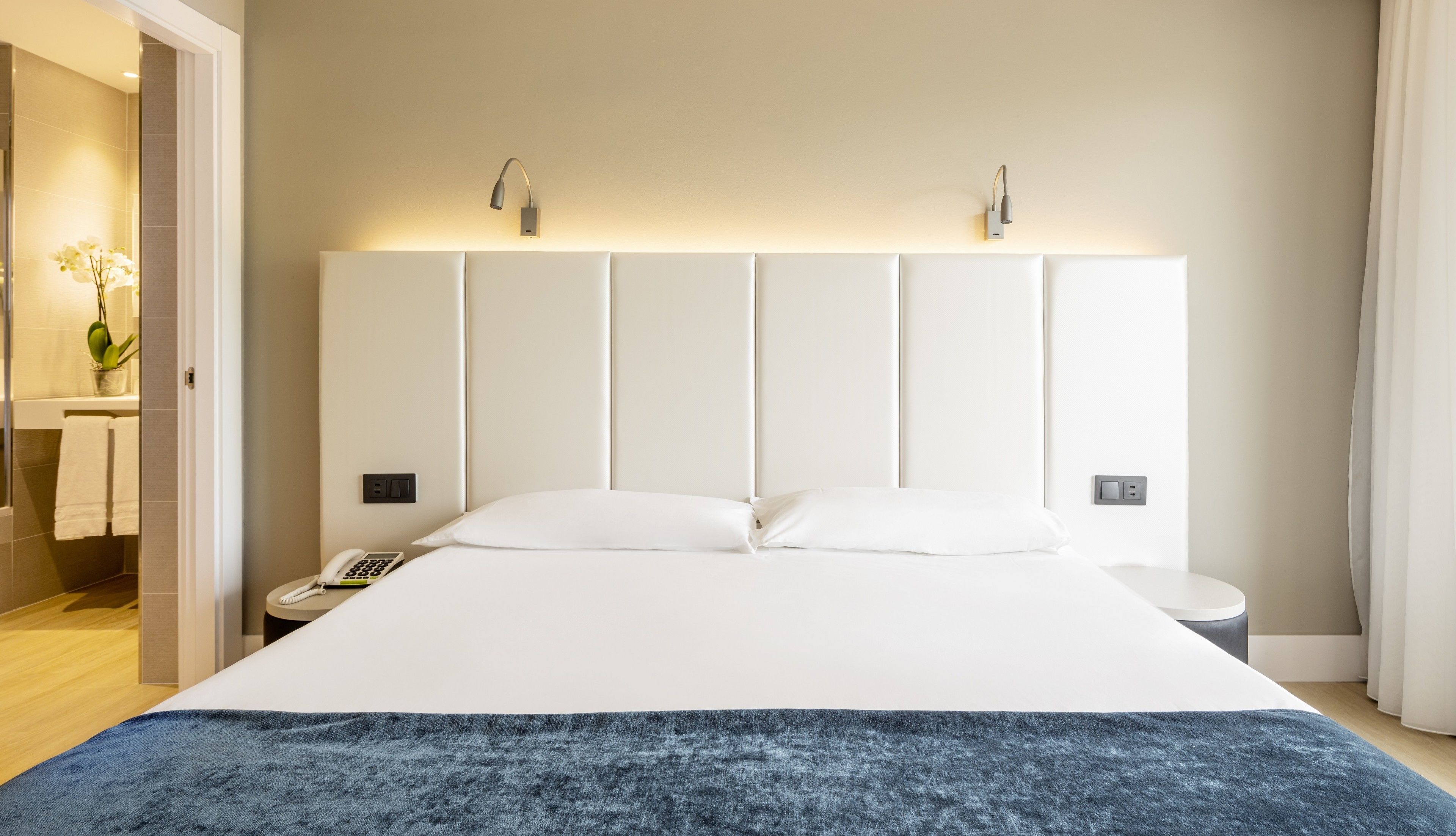 A modern hotel room with a large bed, a bedside table with a phone, and a wall-mounted light fixture.