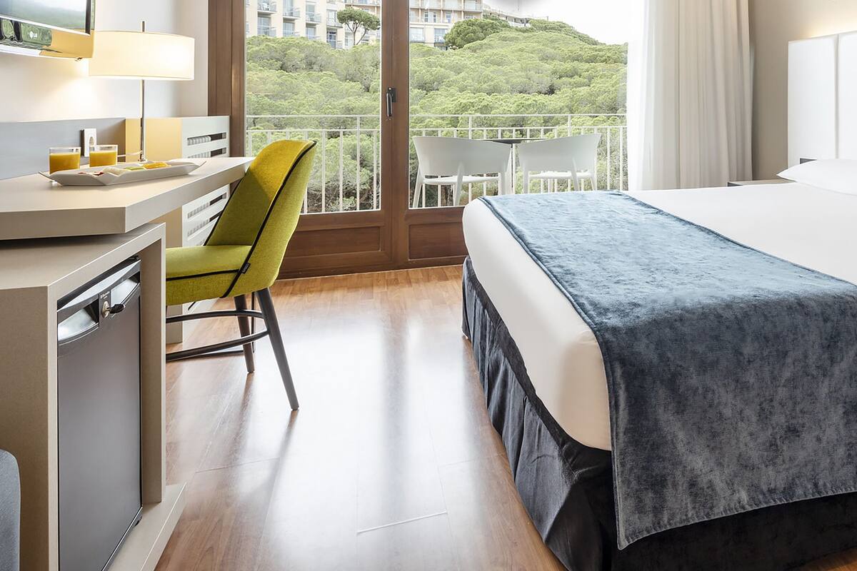 A hotel room with a bed, a desk with a lamp, a chair, and a view of greenery through the window.