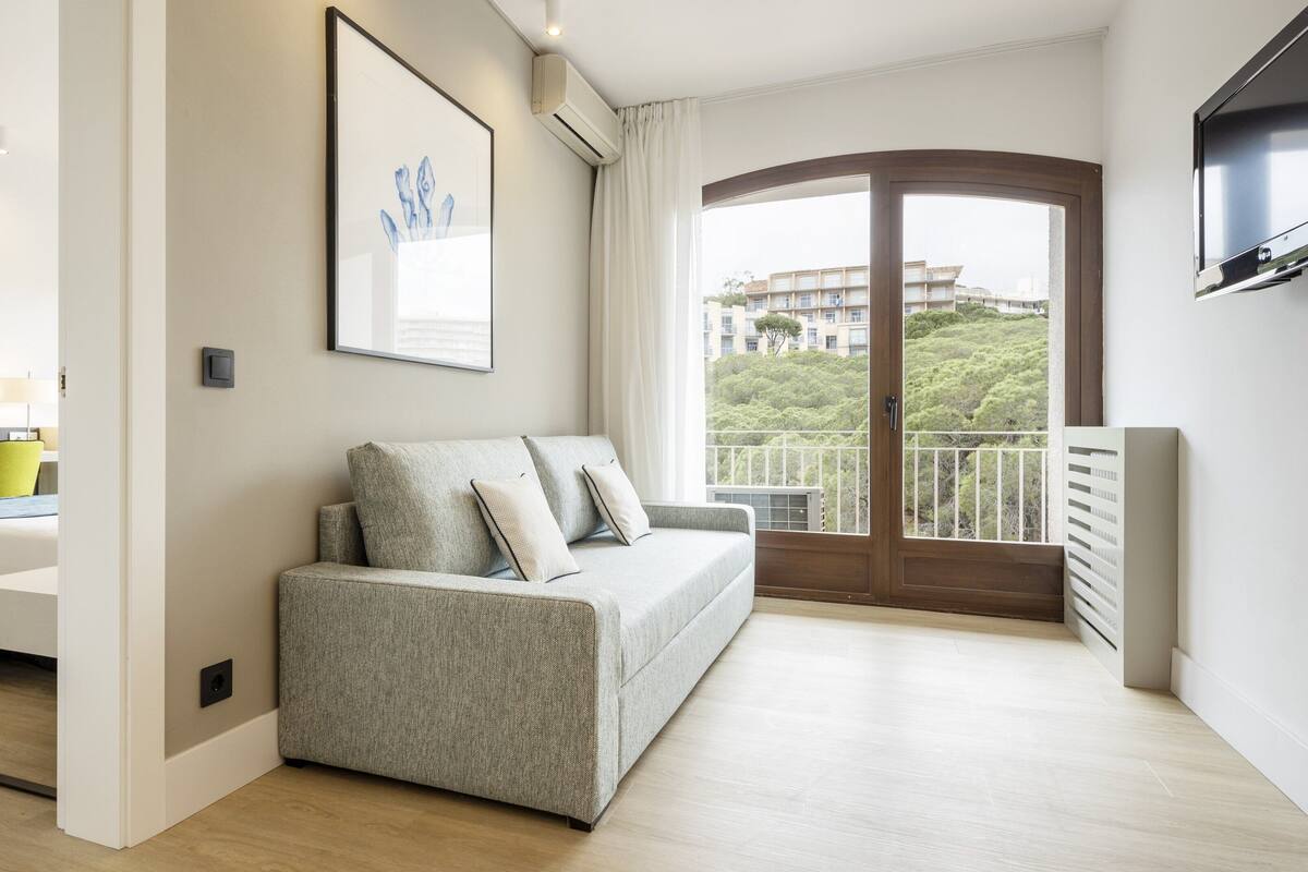 A modern living room with a grey sofa, a flat-screen TV, a dining area, and a balcony with a view of buildings and greenery.
