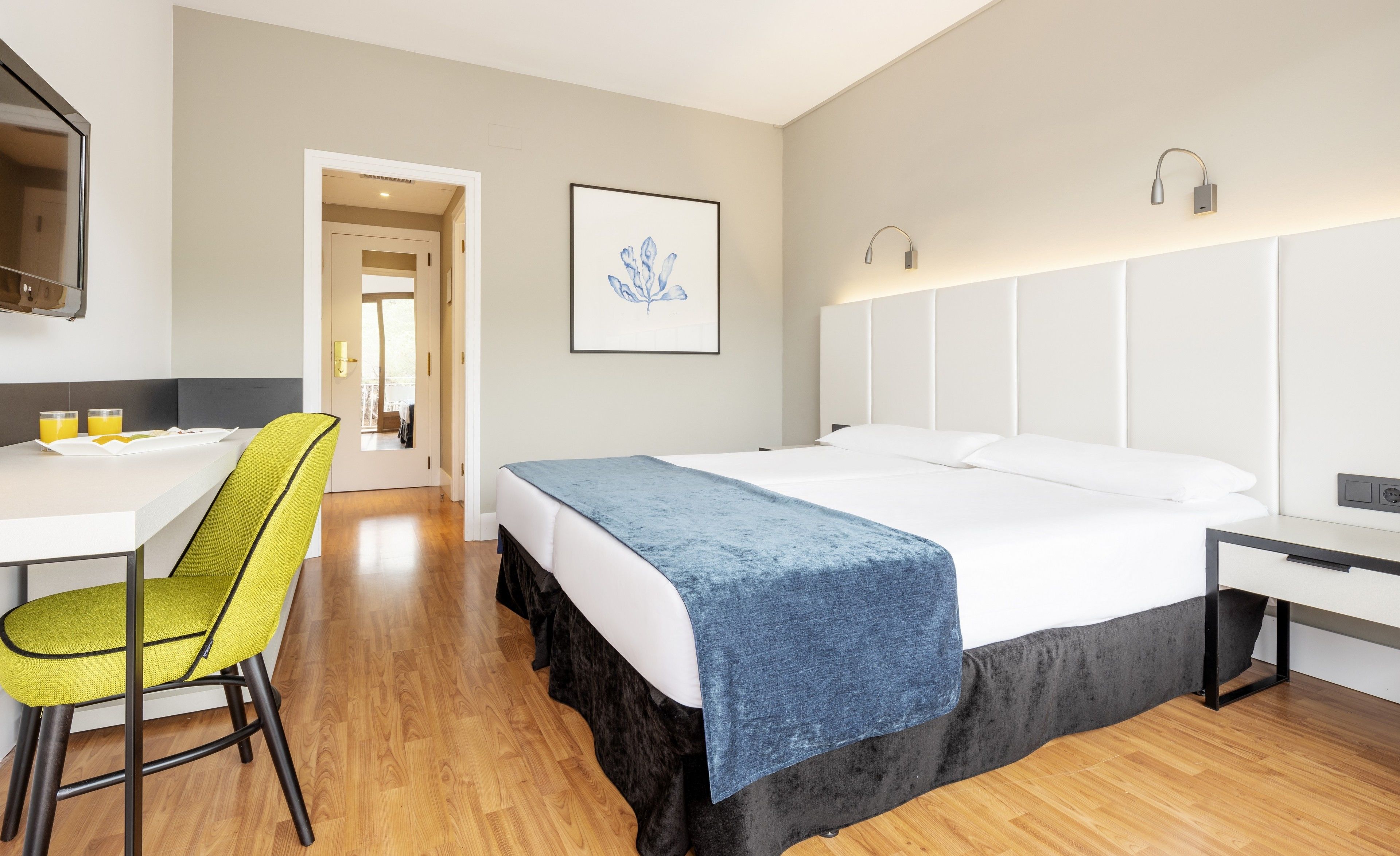A hotel room with a bed, a desk with a chair, a framed artwork, and a door leading to another room.