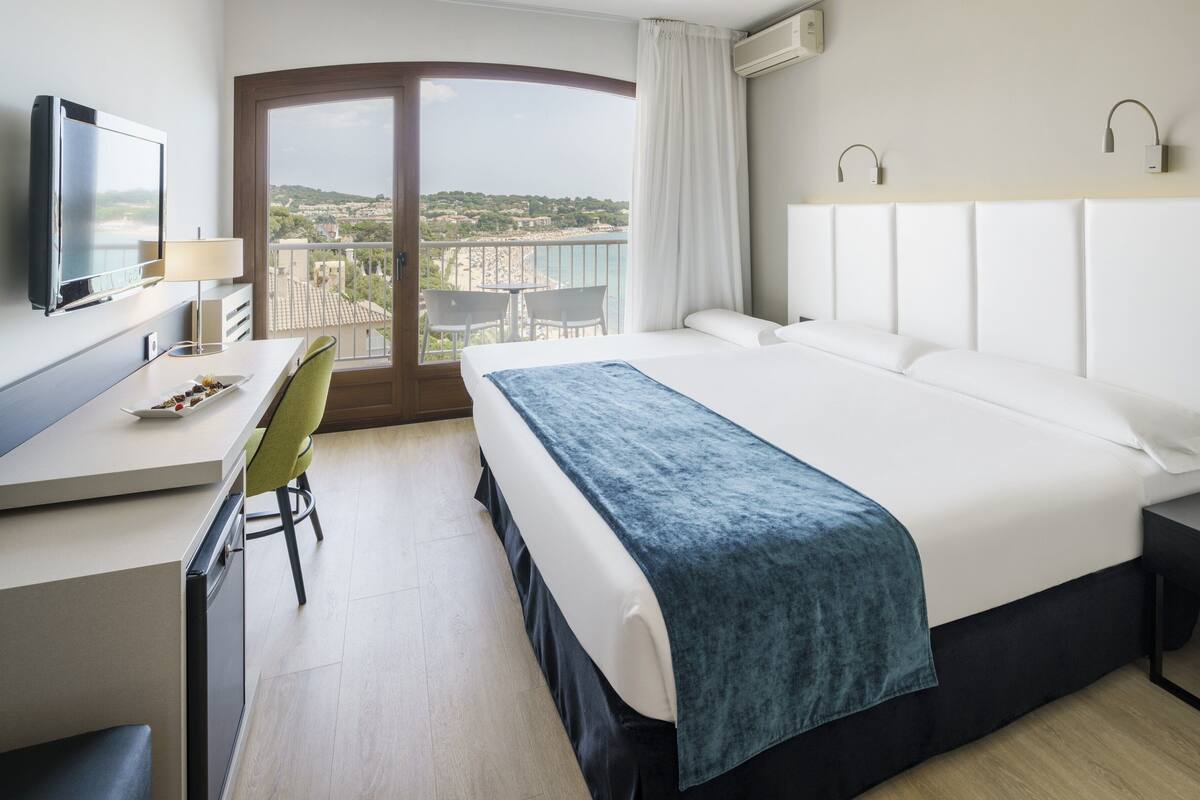 A hotel room with a bed, a desk, a chair, a TV, and a balcony with a view of the city.