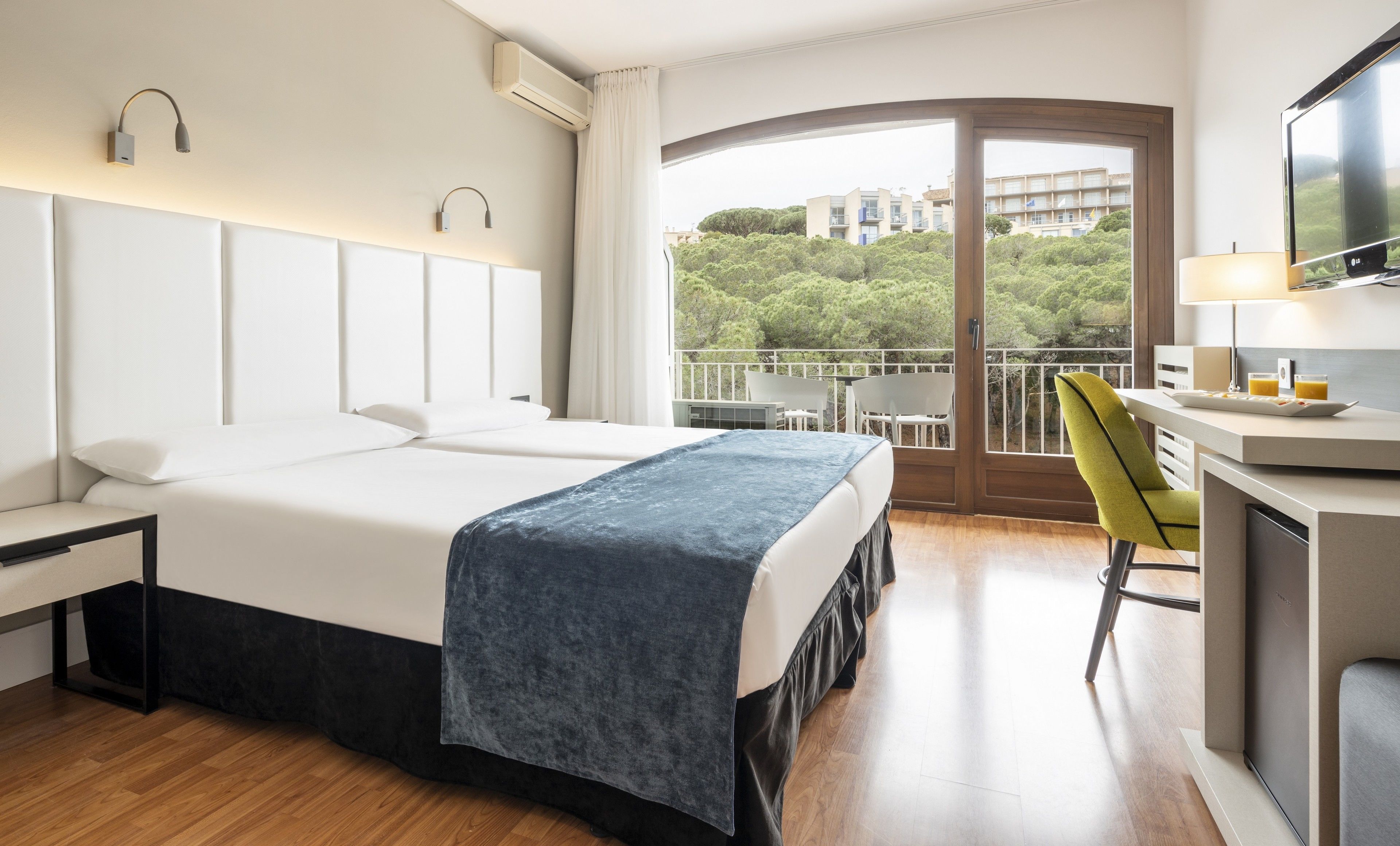 Habitación de hotel con una cama grande, un escritorio con una silla, televisión y balcón con vistas a edificios y árboles.