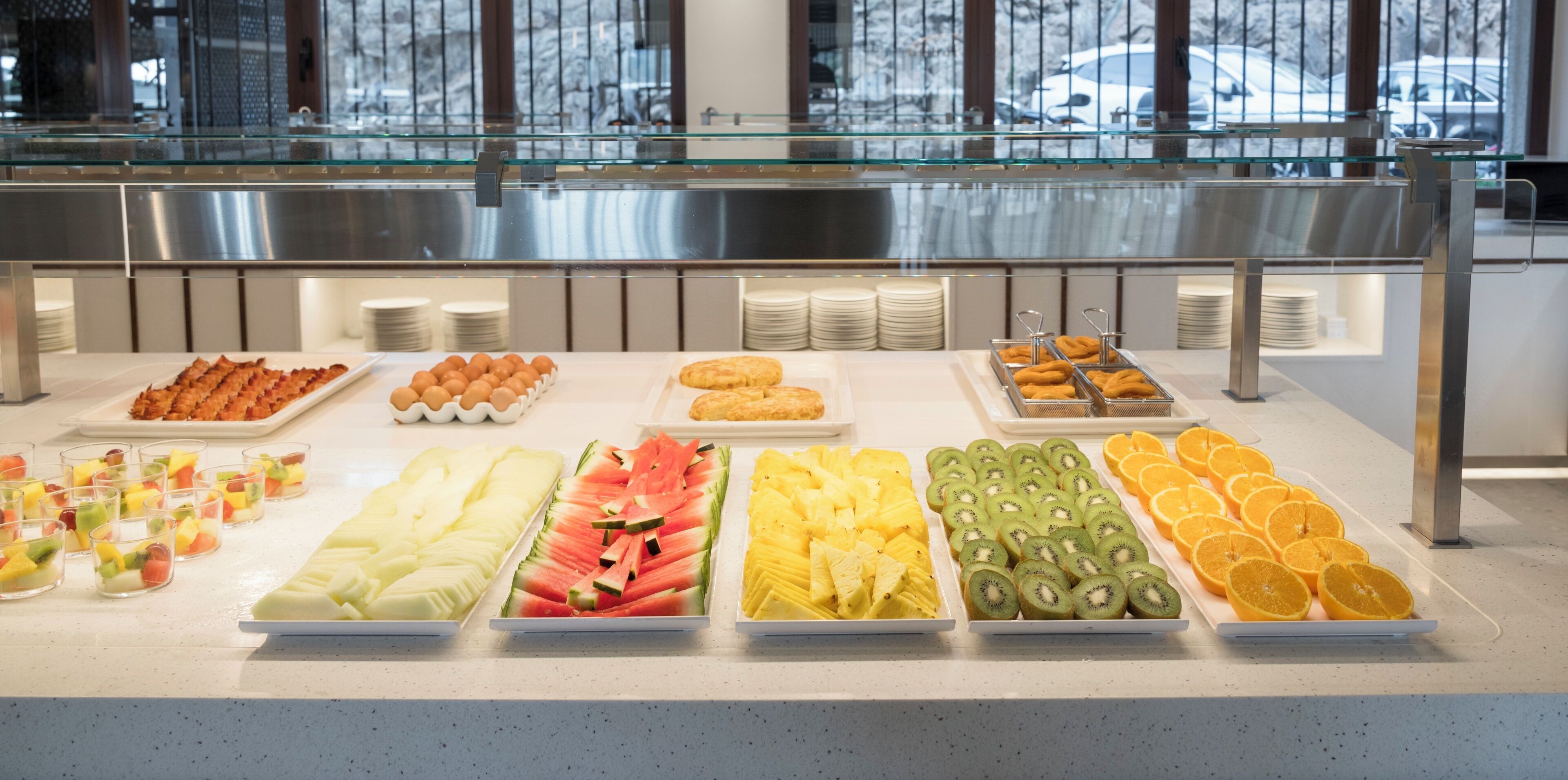 Un bufé con diversas frutas, pasteles y una barandilla de cristal al fondo.